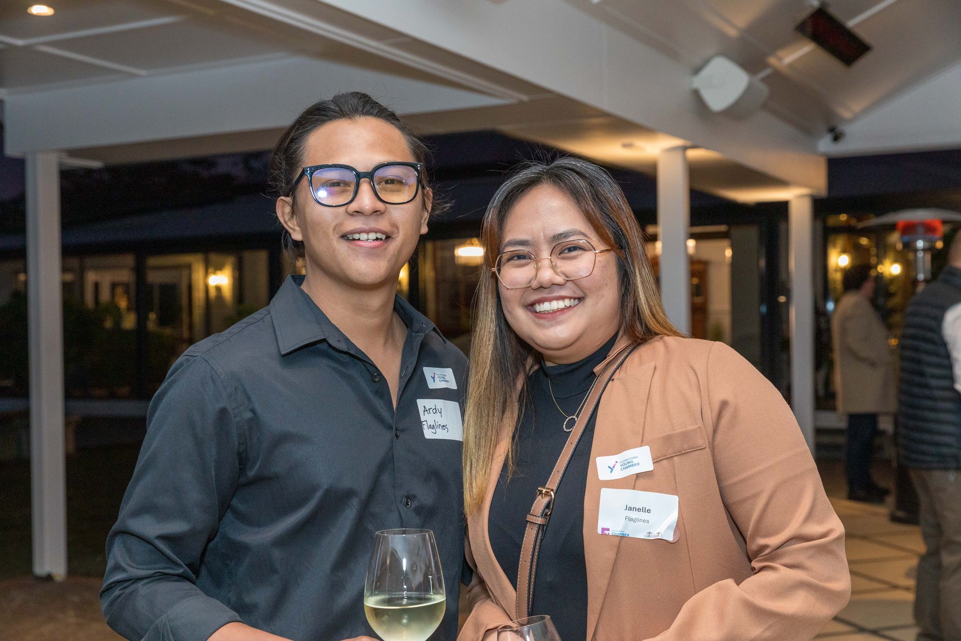 Two smiling people with name tags pose at an event, one holding a glass of wine. They stand indoors, with soft lighting and visible guests in the background.