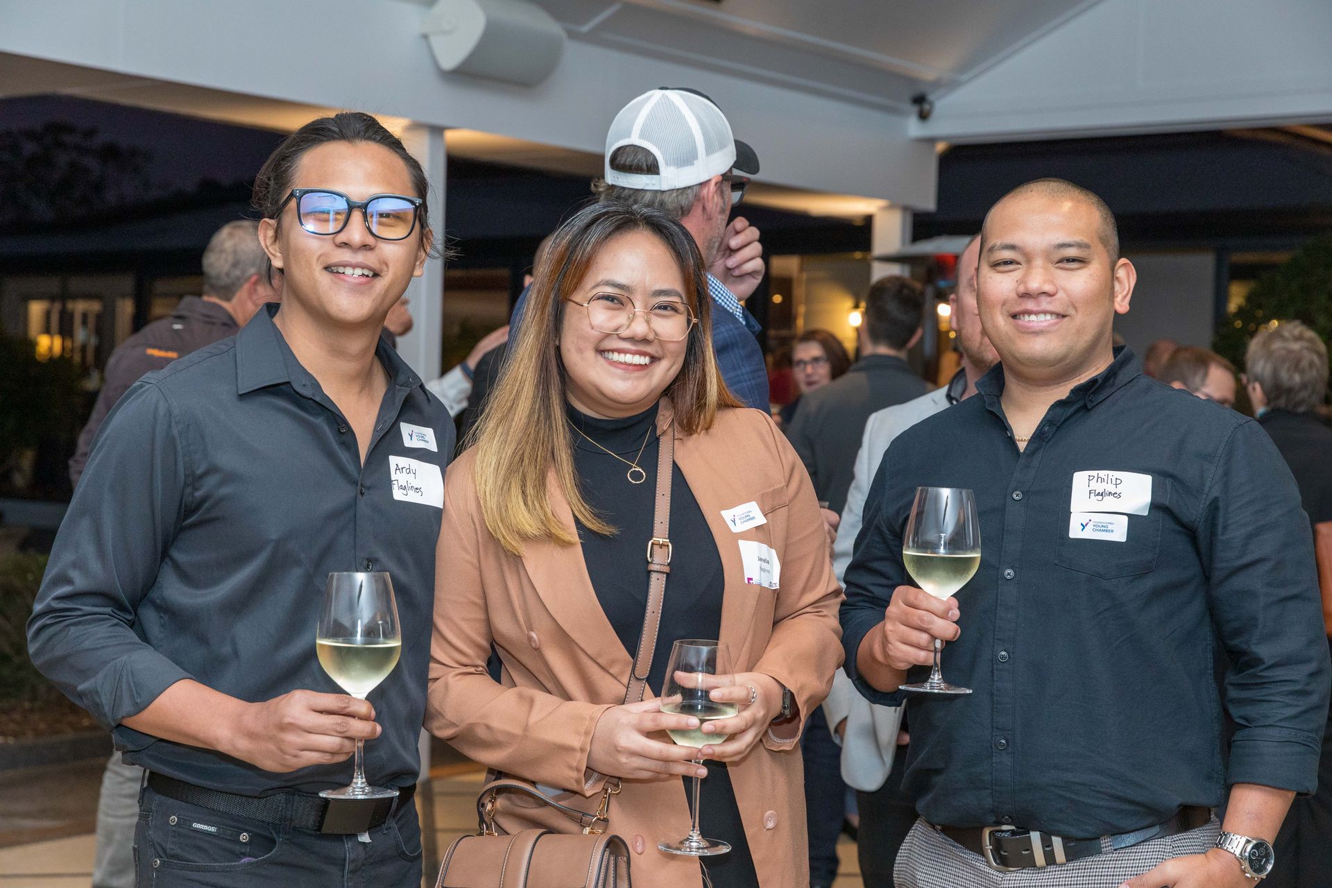 Three people smiling, holding wine glasses, at an outdoor event. A woman in a tan coat stands between two men, all wearing name tags.