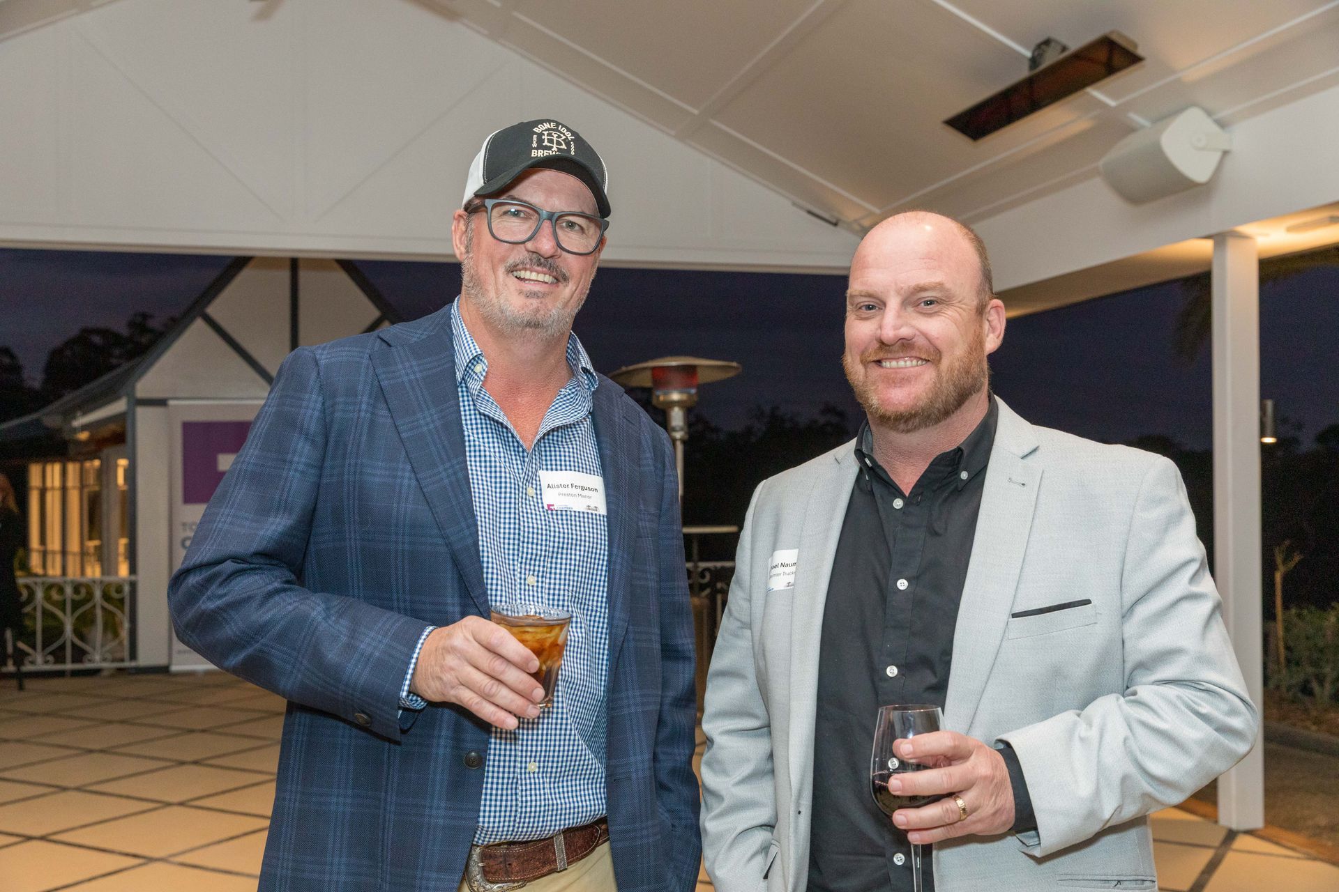 Two men at an outdoor event, smiling and holding drinks. One wears a blue blazer and a baseball cap, the other a grey blazer.