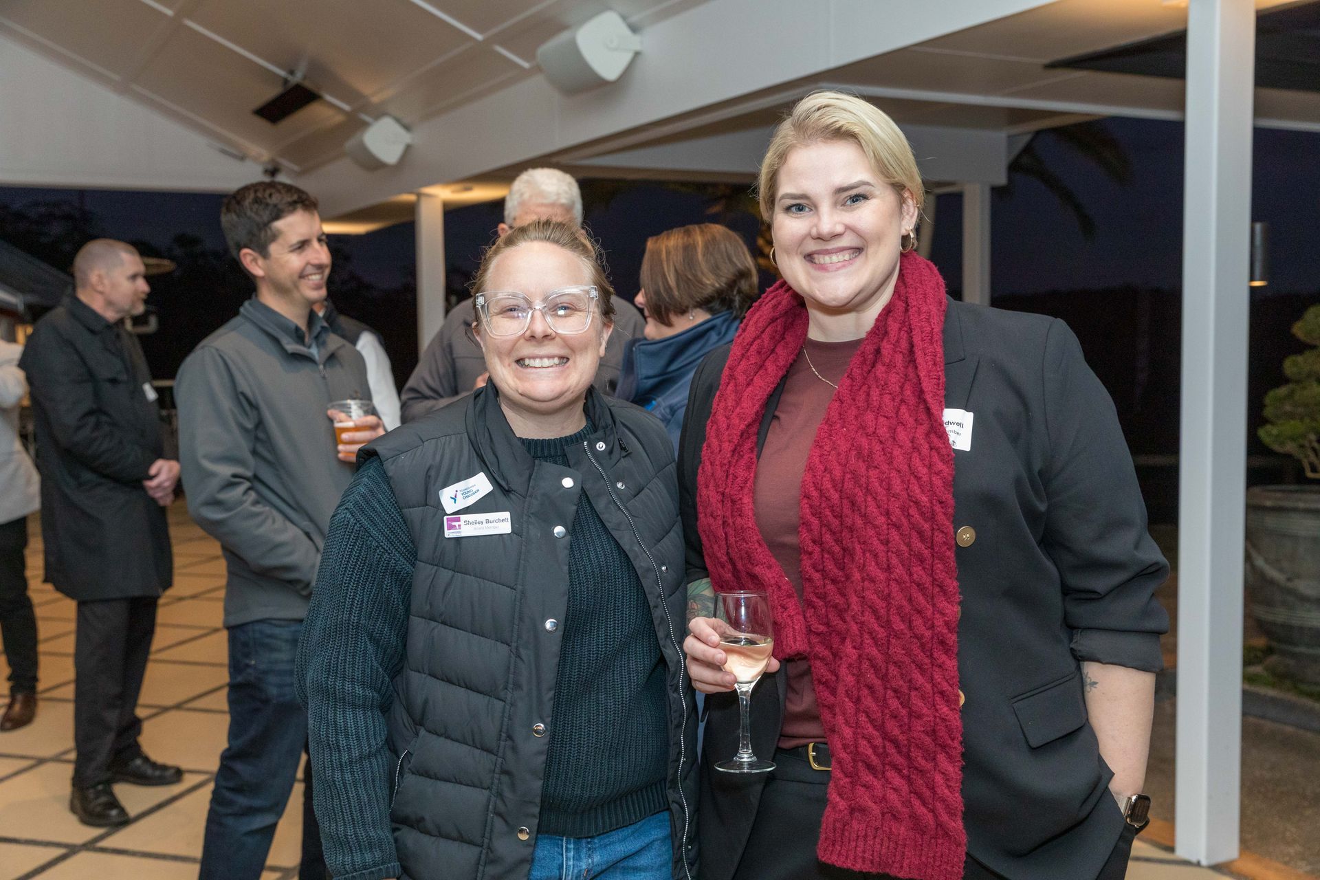Two women smiling and posing at an outdoor gathering. One wears a black blazer and red scarf, the other a vest and glasses.