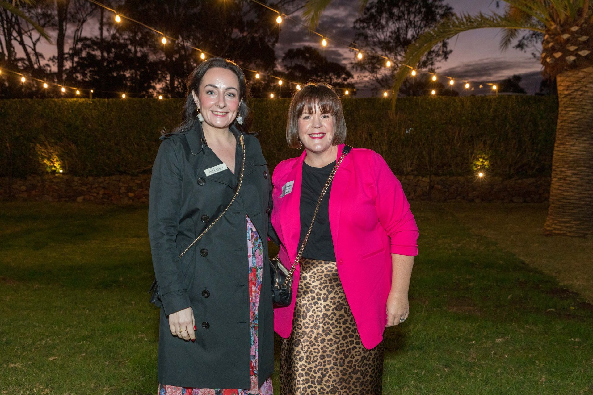 Two women smile, posing outdoors at dusk. One wears a black coat and the other a pink blazer and leopard-print skirt. String lights illuminate the background.