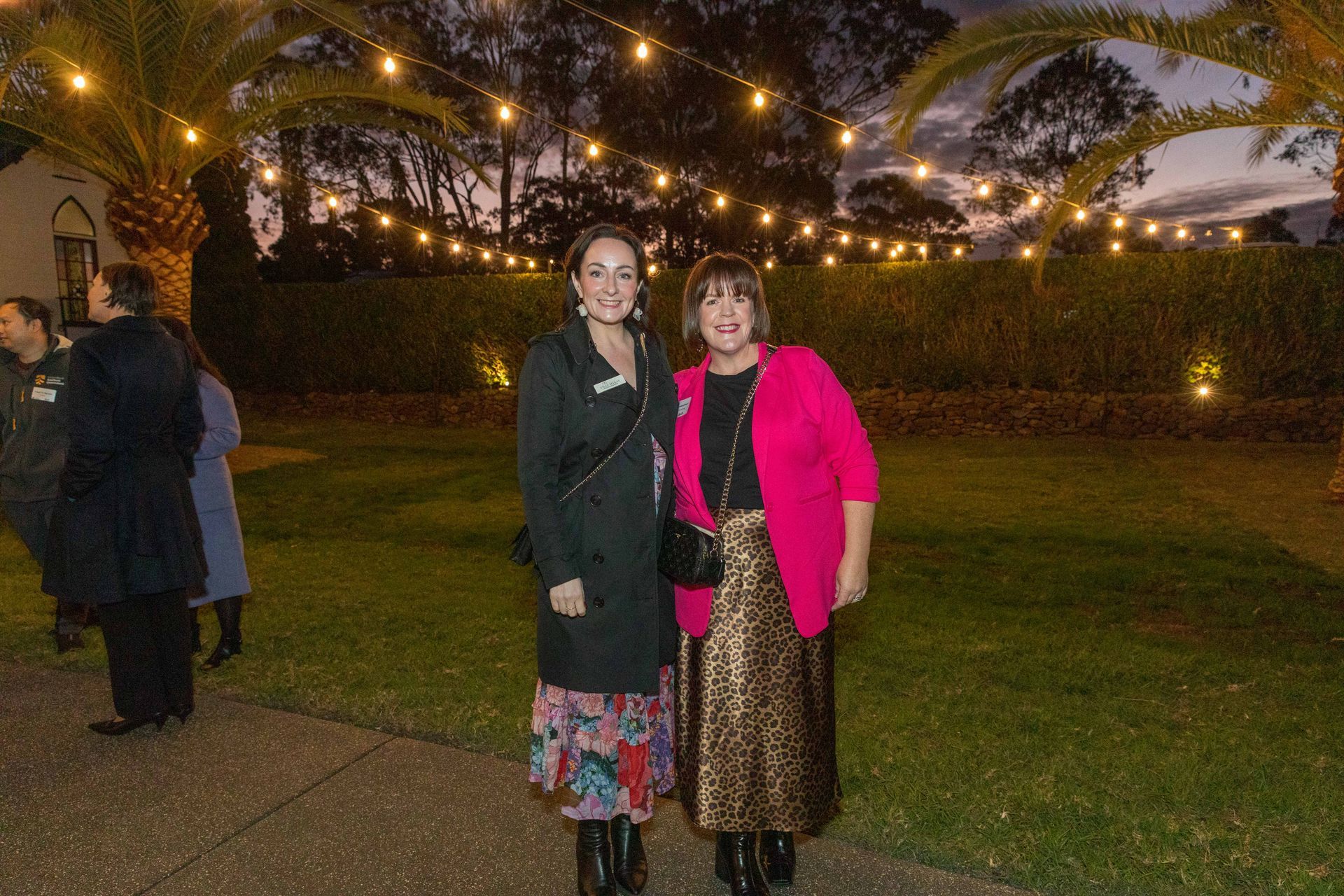 Two women stand outside at dusk. One wears a black coat and floral skirt, the other a pink jacket and gold skirt, smiling under string lights.