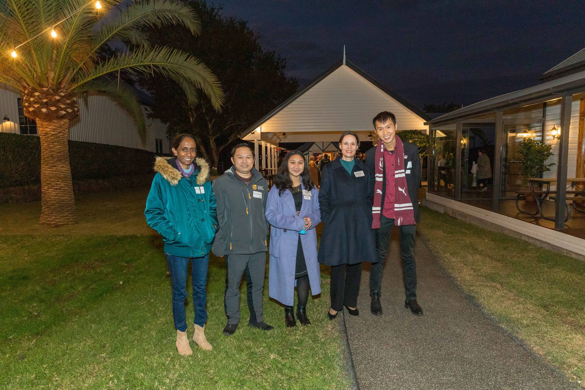 Five people stand outside a building at dusk. The group is diverse in clothing, on a grassy area with string lights overhead.
