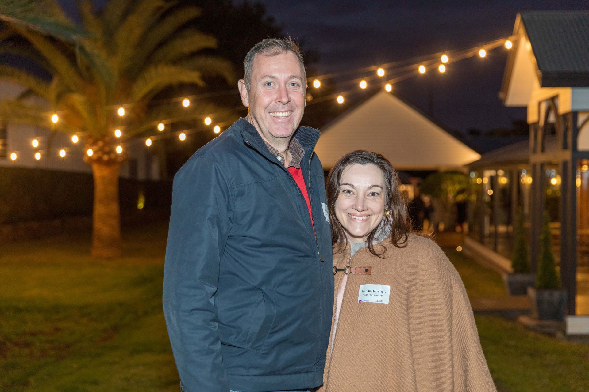 Man and woman smiling, standing outside at night, strung lights in the background.