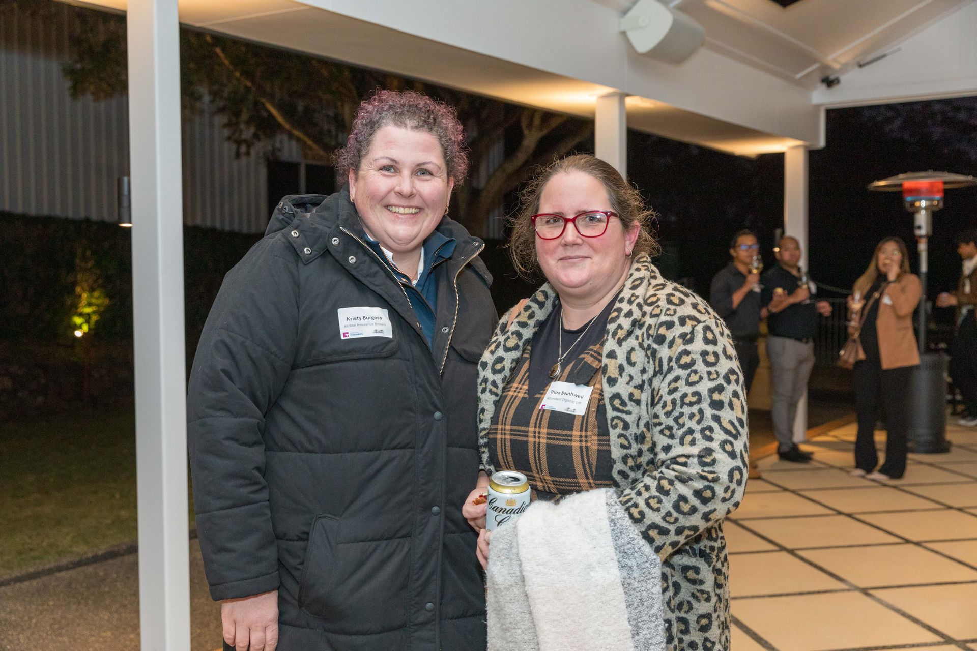 Two women smiling at an outdoor event. The woman on the left wears a black coat; the woman on the right wears a leopard print coat.