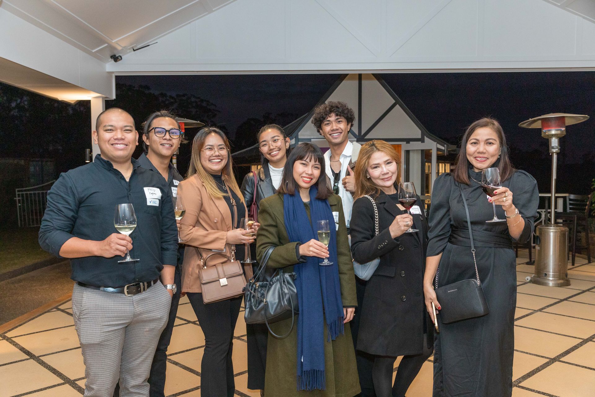 Group of diverse people at an outdoor event, holding wine glasses and smiling. They are in a patio setting at dusk.