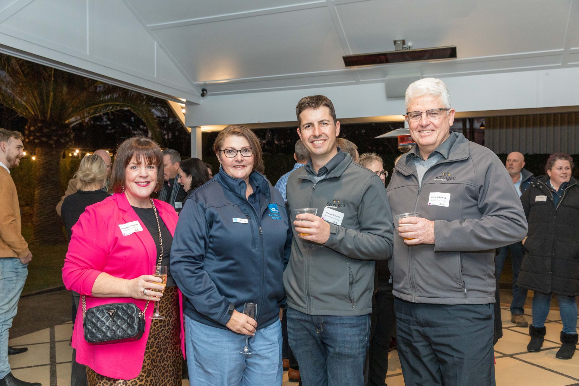 Four people at an outdoor evening event. Two women and two men smile, holding drinks. Setting is a covered patio with soft lighting.