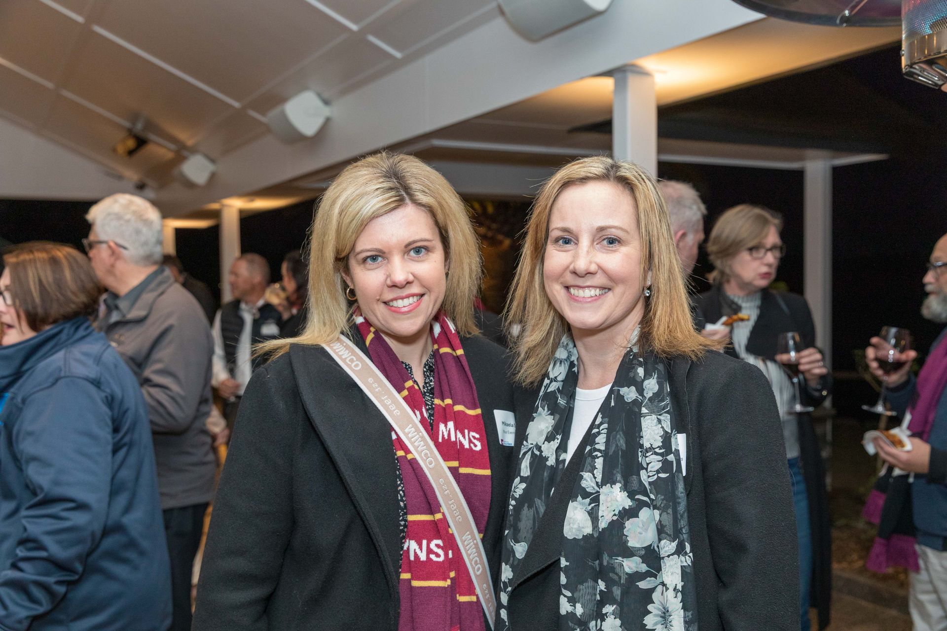 Two women smiling at an event, posing for a photo. One wears a maroon scarf, the other a patterned black and white scarf. Other guests stand in the background.