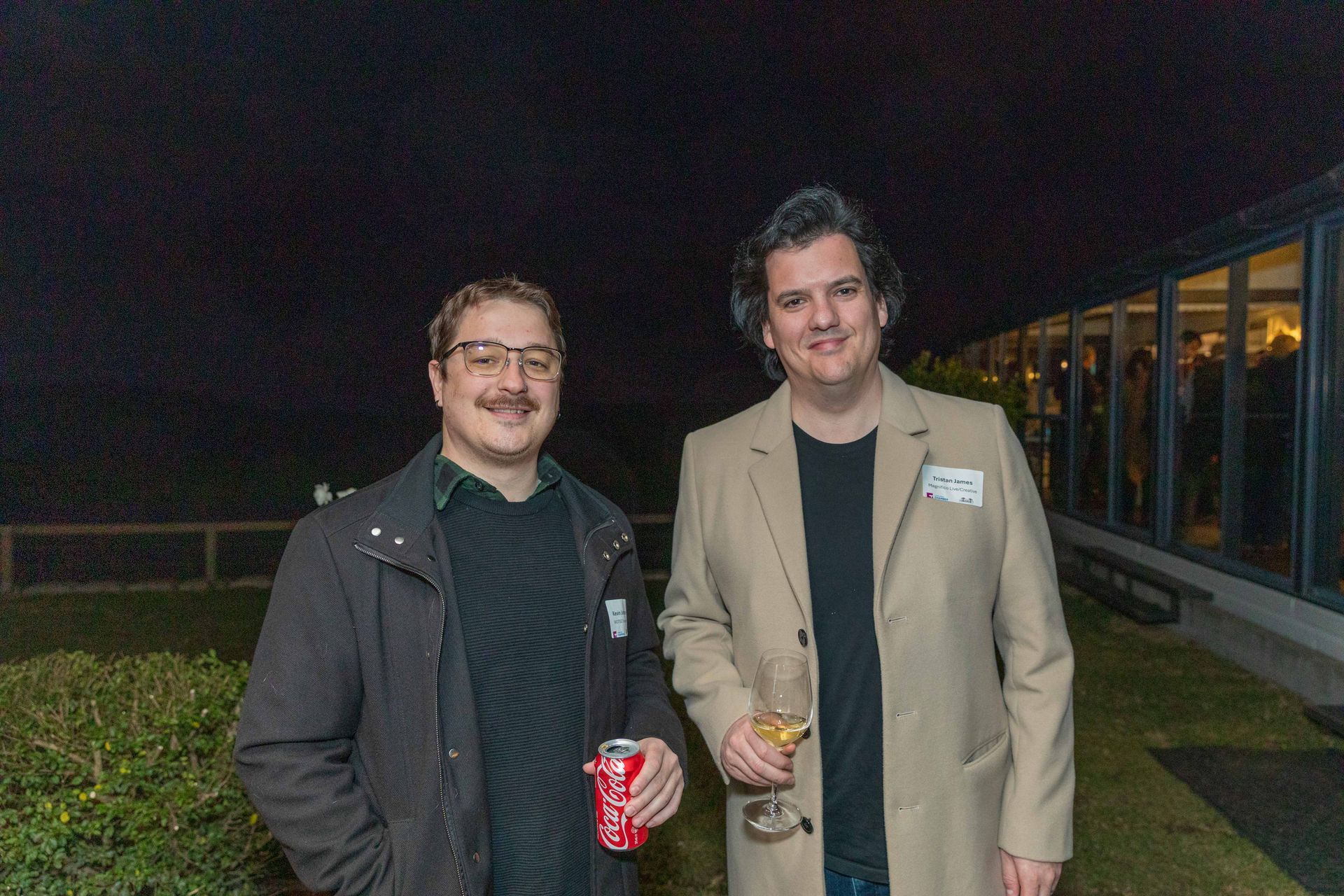 Two men smiling, one holding a drink and the other a can, outdoors at night. They're in front of a building with lights on, and a dark sky is above.