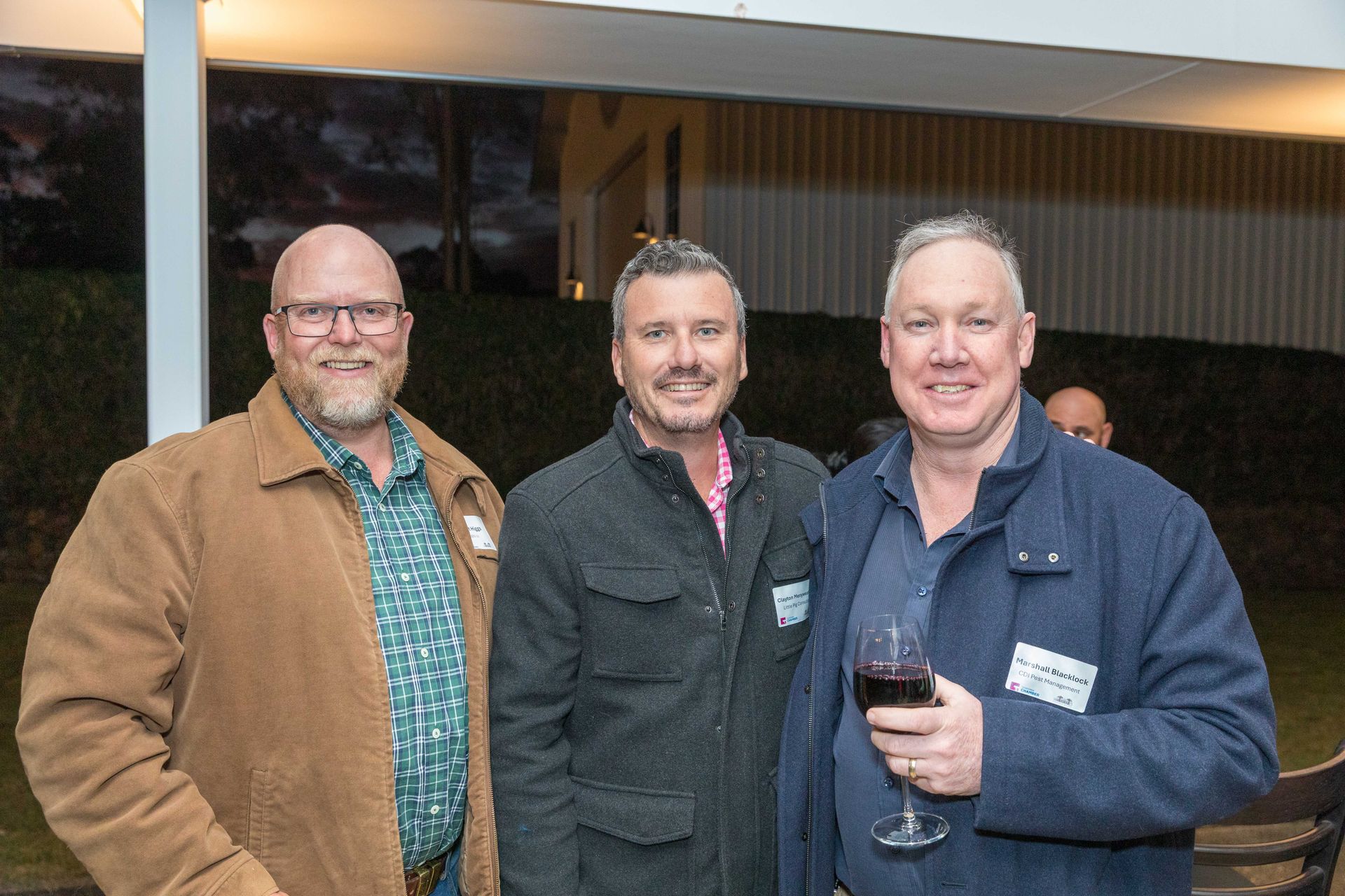 Three men at an outdoor gathering, smiling. One holds a glass of red wine. They are wearing casual jackets.