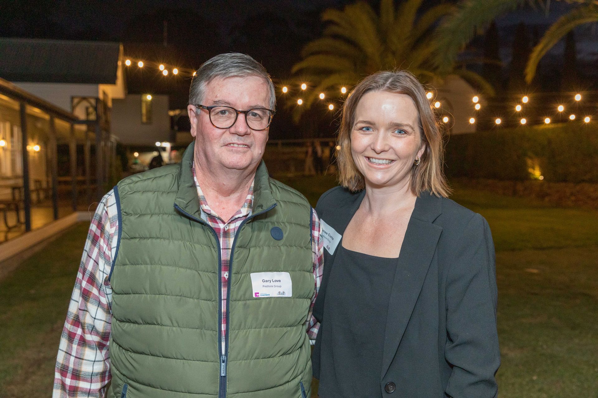Man and woman smiling, posing for a photo outdoors at night. Man in glasses, vest, and plaid shirt; woman in a black blazer. Lit-up background.