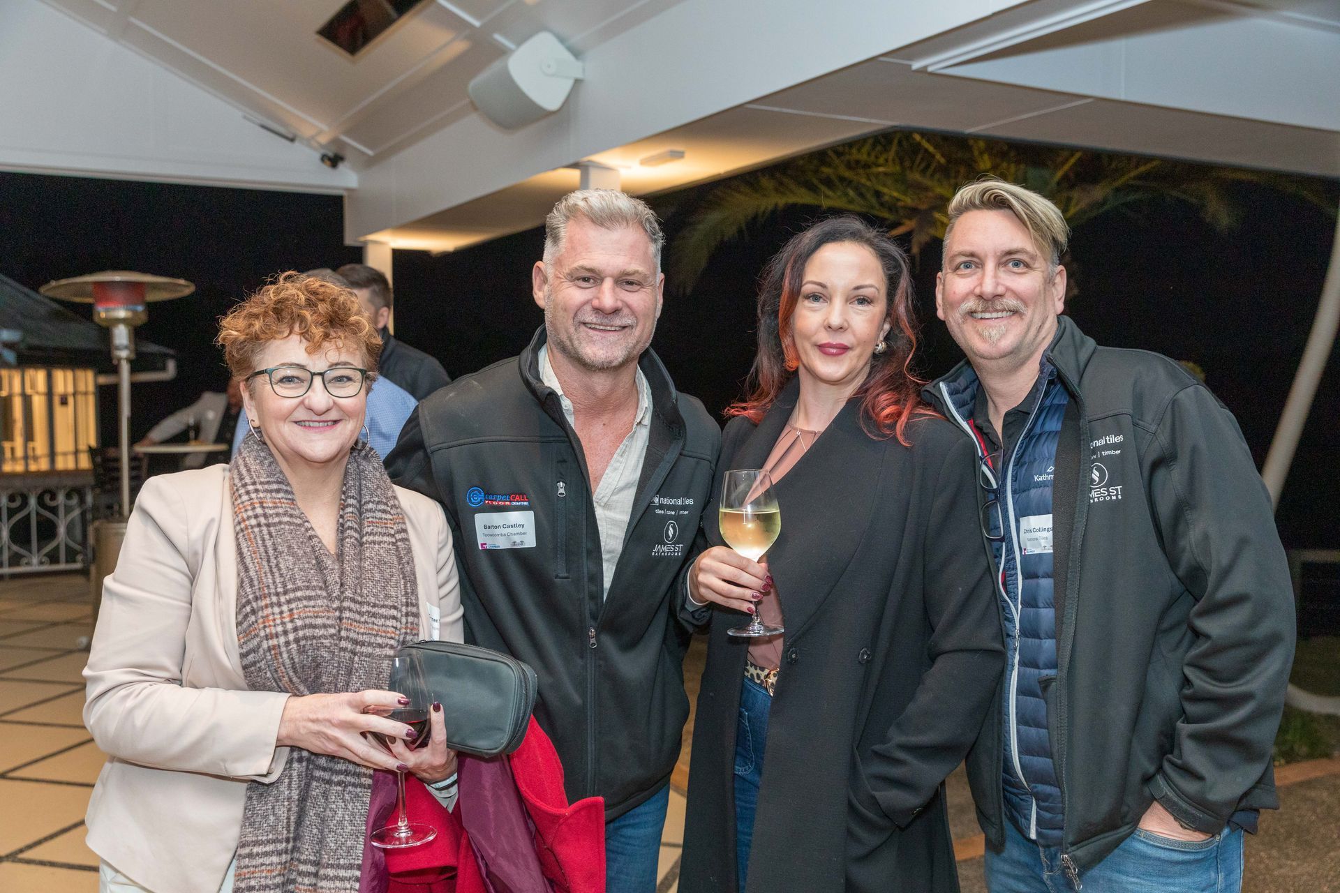 Four people smiling and posing together outside at night. A woman in a blazer and glasses stands with a man in a black jacket, and another couple to the right.