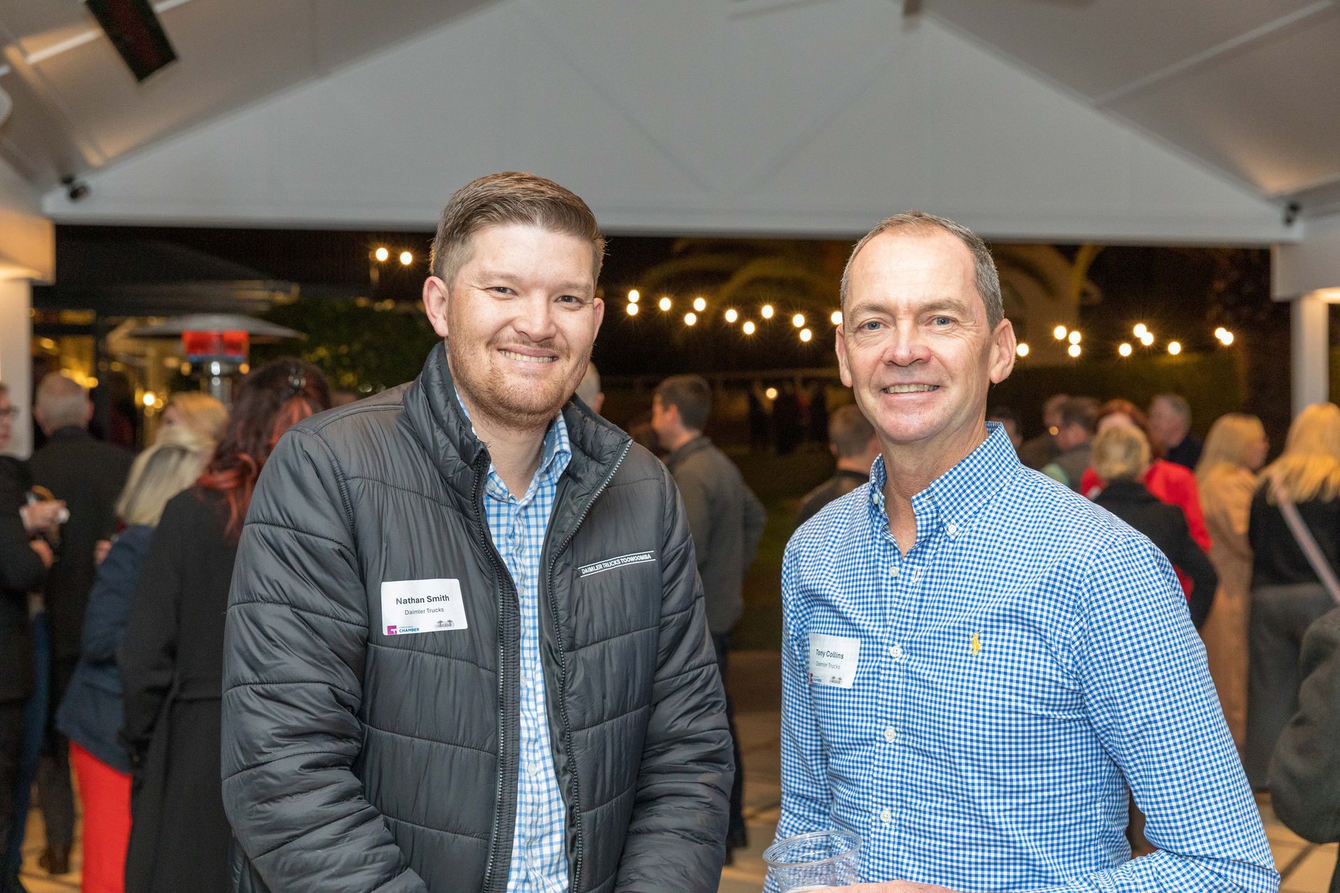 Two men smiling, posing at an outdoor event. The man on the left wears a black jacket. The other man wears a blue checkered shirt.