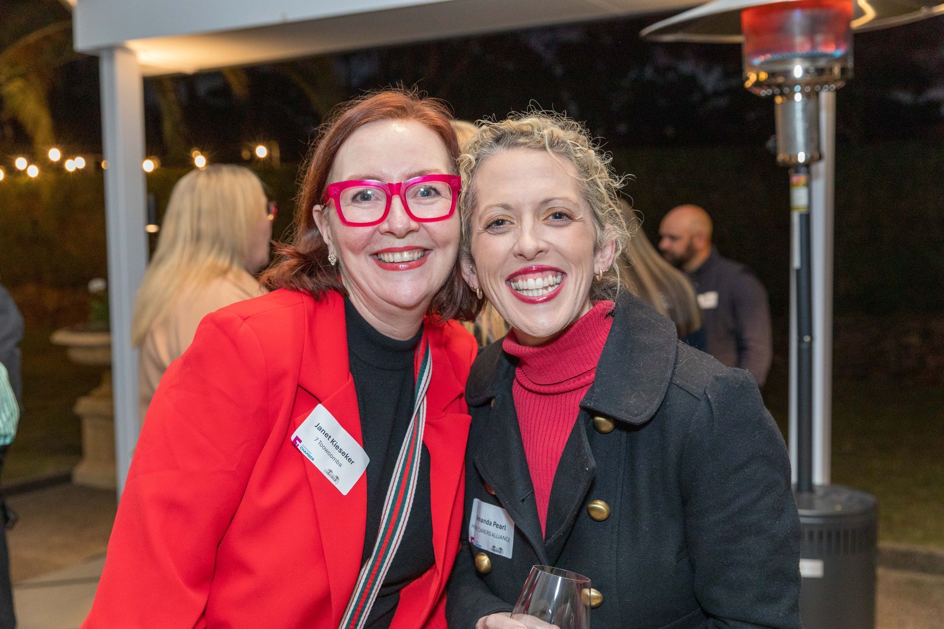 Two smiling women pose for a photo outdoors. One wears a red blazer and pink glasses. The other has a black coat and red turtleneck.