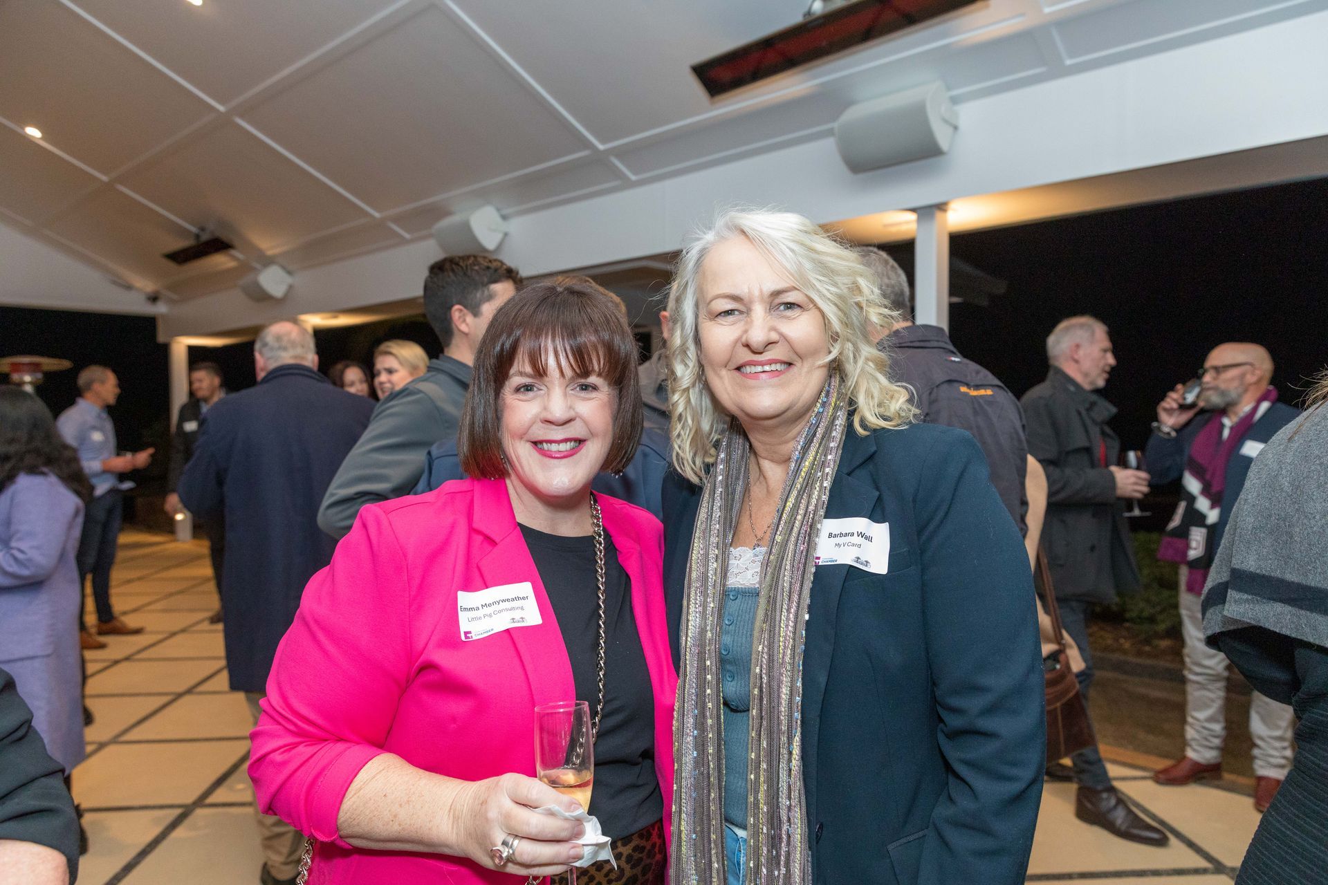 Two women smiling at an outdoor event. One wears a pink jacket, the other a navy jacket with a scarf. Guests mingle in the background.