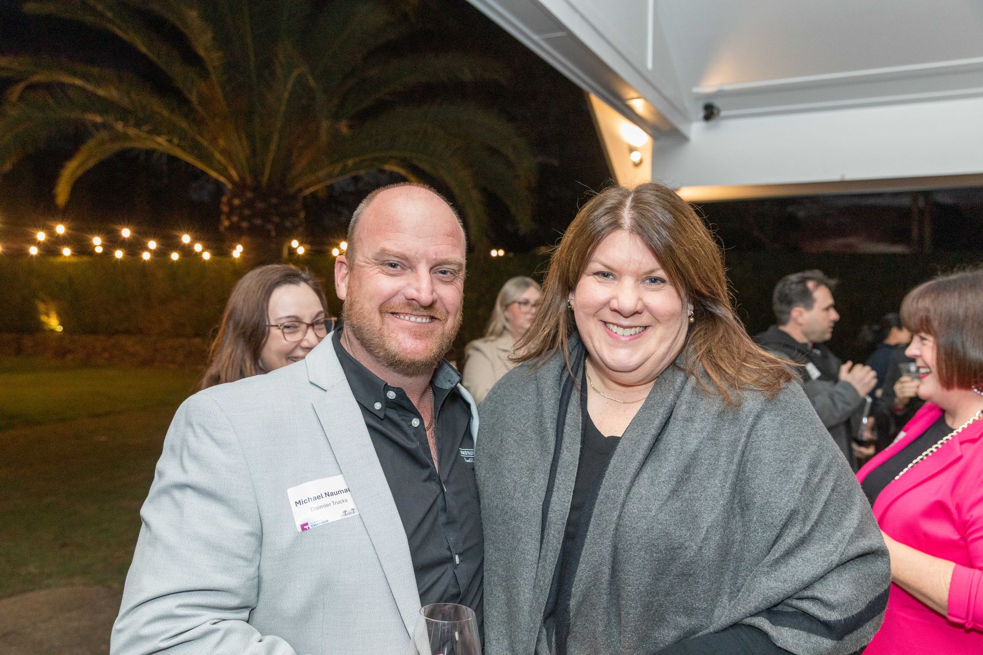 A man and a woman smile at the camera at an outdoor event. The man wears a light grey blazer, the woman a dark grey shawl.