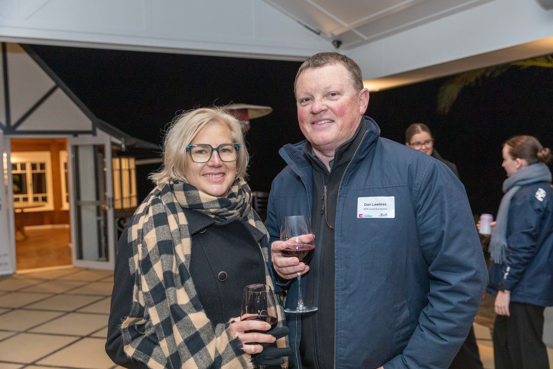 Two people, a woman and a man, smiling and holding drinks outside at night. The woman wears a scarf and glasses, the man a jacket.