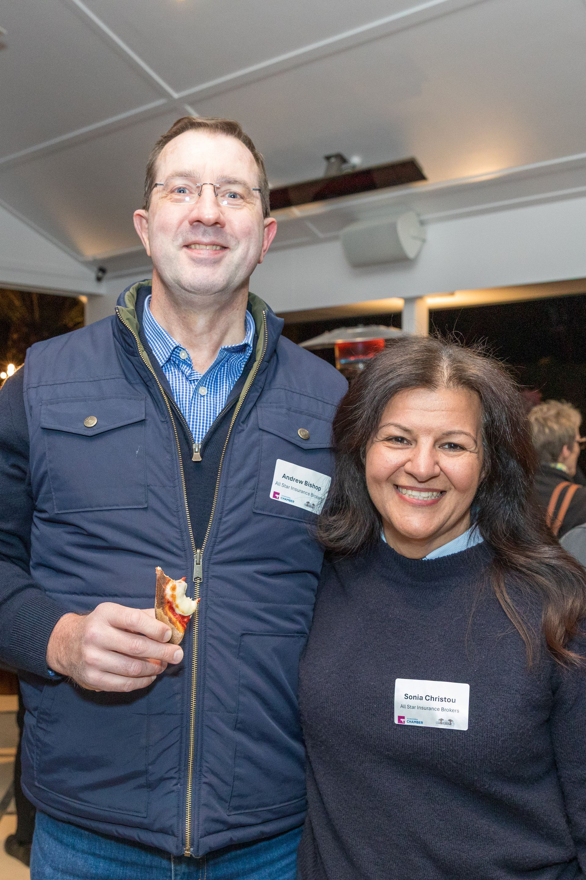 A man and woman smile for the camera. The man holds a small toy and wears a blue vest. The woman wears a blue sweater. They are indoors.