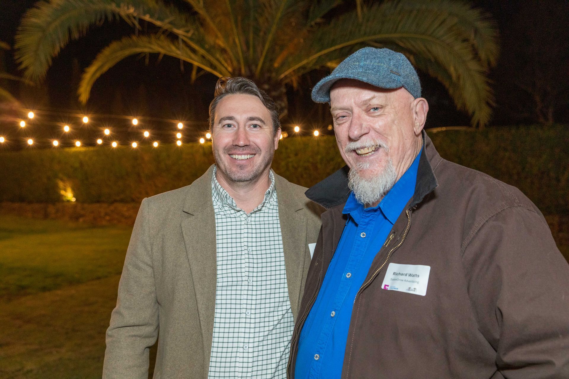 Two men smiling, posing outdoors at night, with string lights and a palm tree in the background.