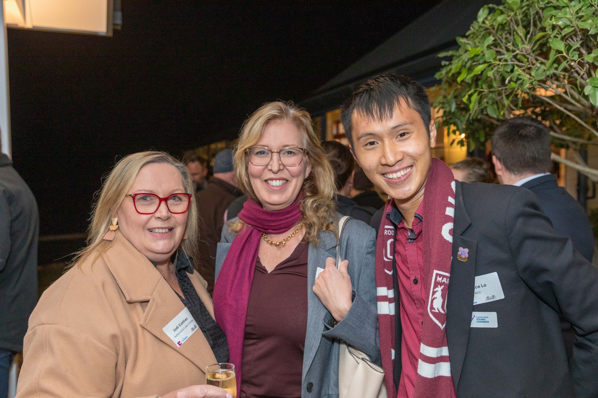 Three people smiling for a photo outside at night. A woman on the left wears glasses and a tan coat, the woman in the center a maroon scarf, and the man on the right a striped scarf.
