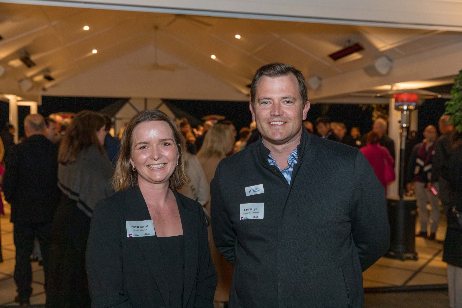 Woman and man smiling at an outdoor event, both wearing name tags. A crowd of people are in the background.