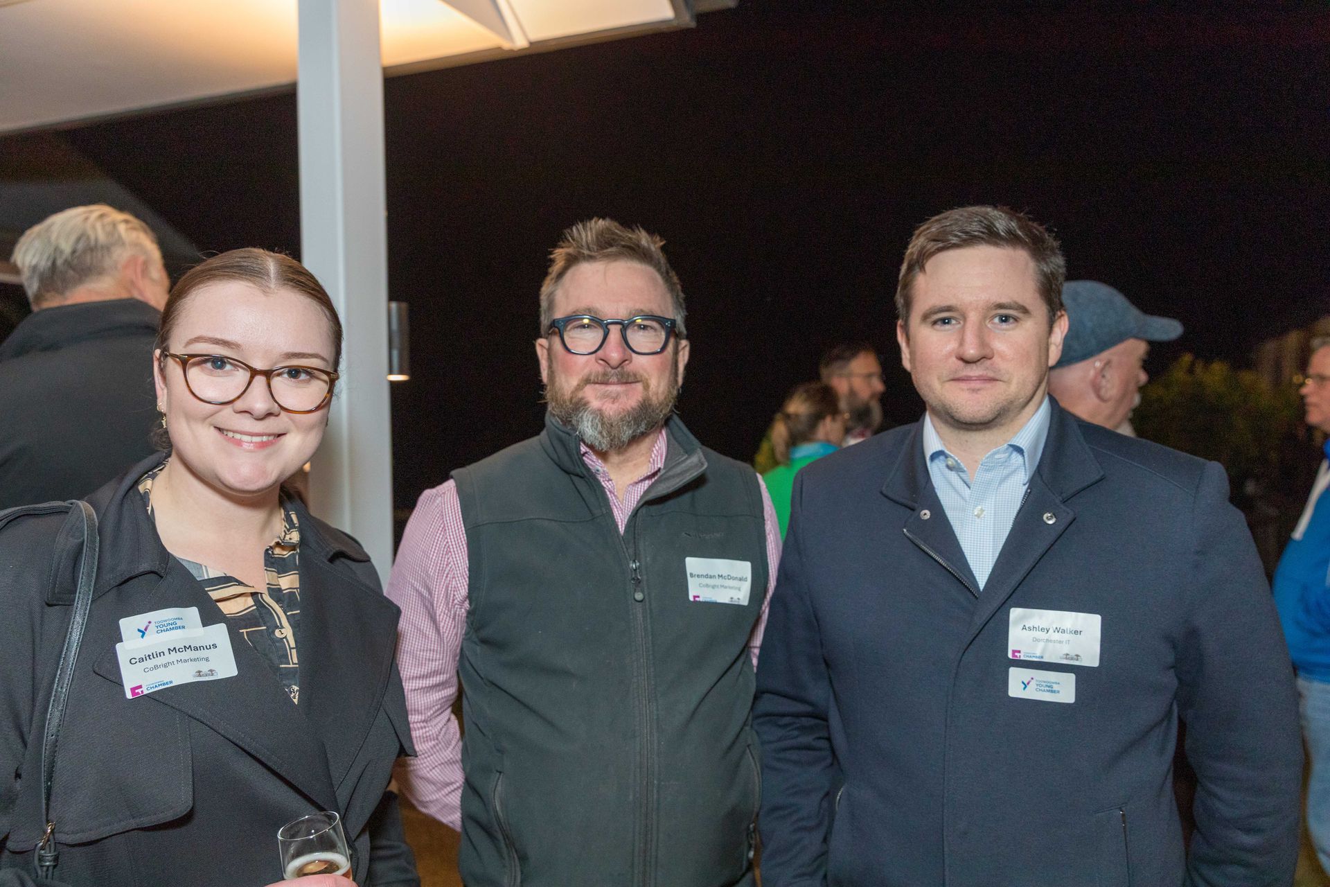 Three people, two men and a woman, at an outdoor evening event. They're wearing name tags and smiling.