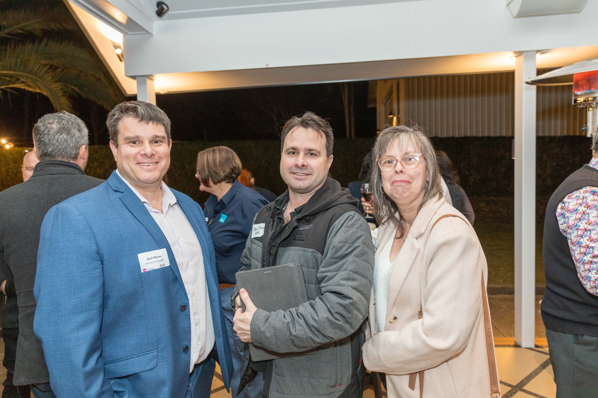 Three people smiling at an outdoor event. A man in a blue blazer stands next to a man holding a tablet, and a woman in a tan coat.