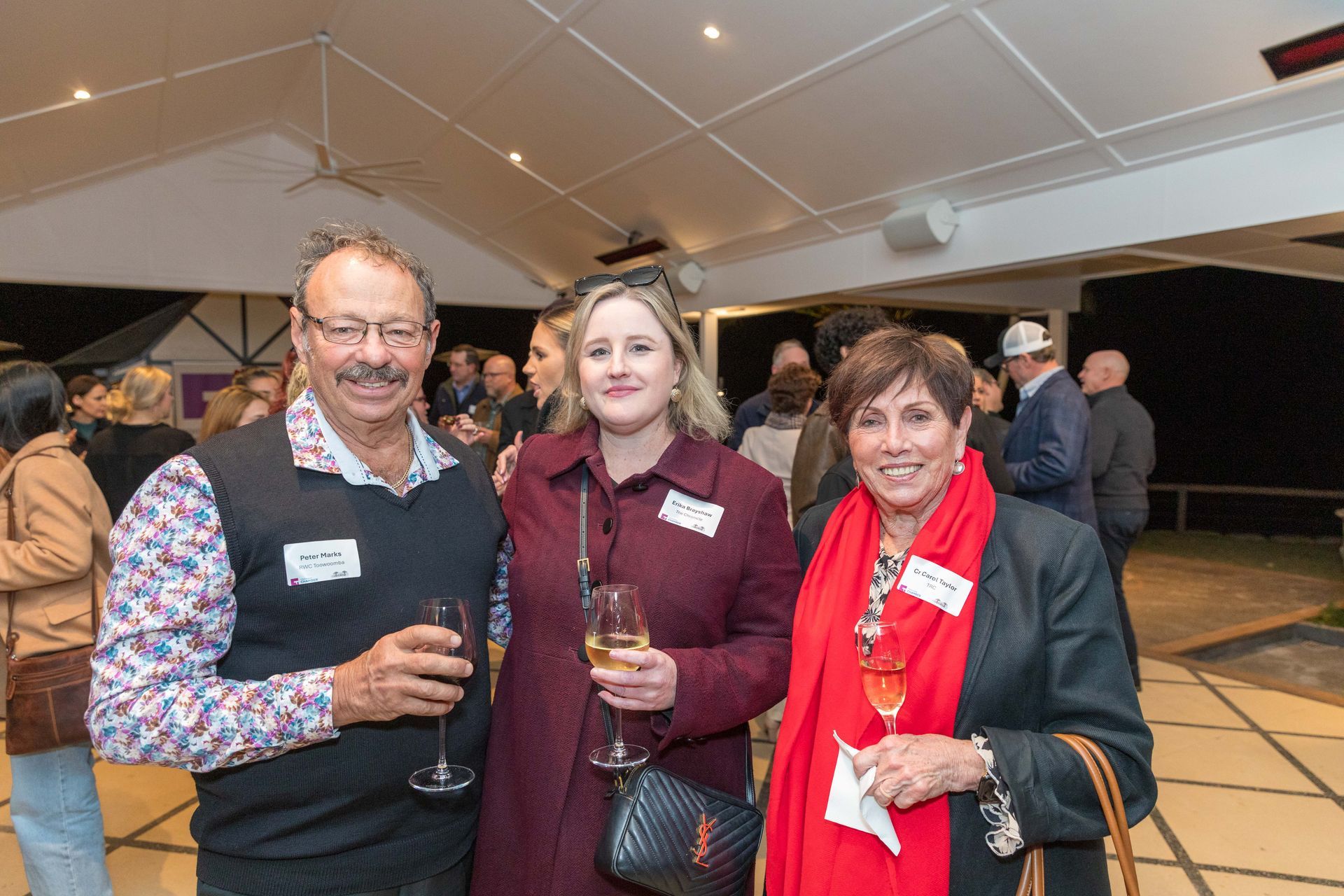 Three people smiling and holding drinks at an event. The man wears a vest, the woman in the middle wears a coat, and the other woman has a red scarf.