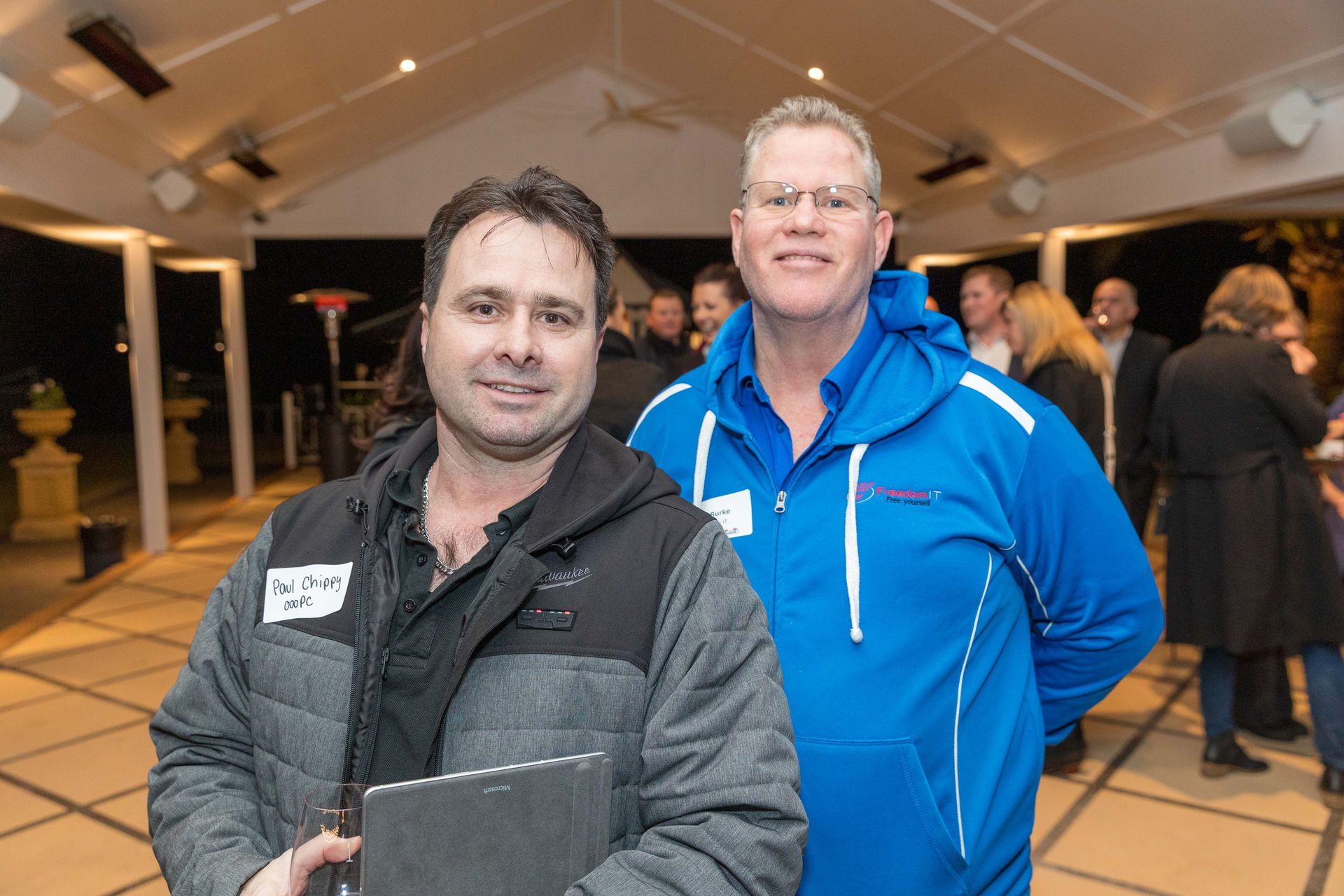 Two men smiling at an outdoor event, one in a blue jacket, the other in a grey vest, holding a tablet.