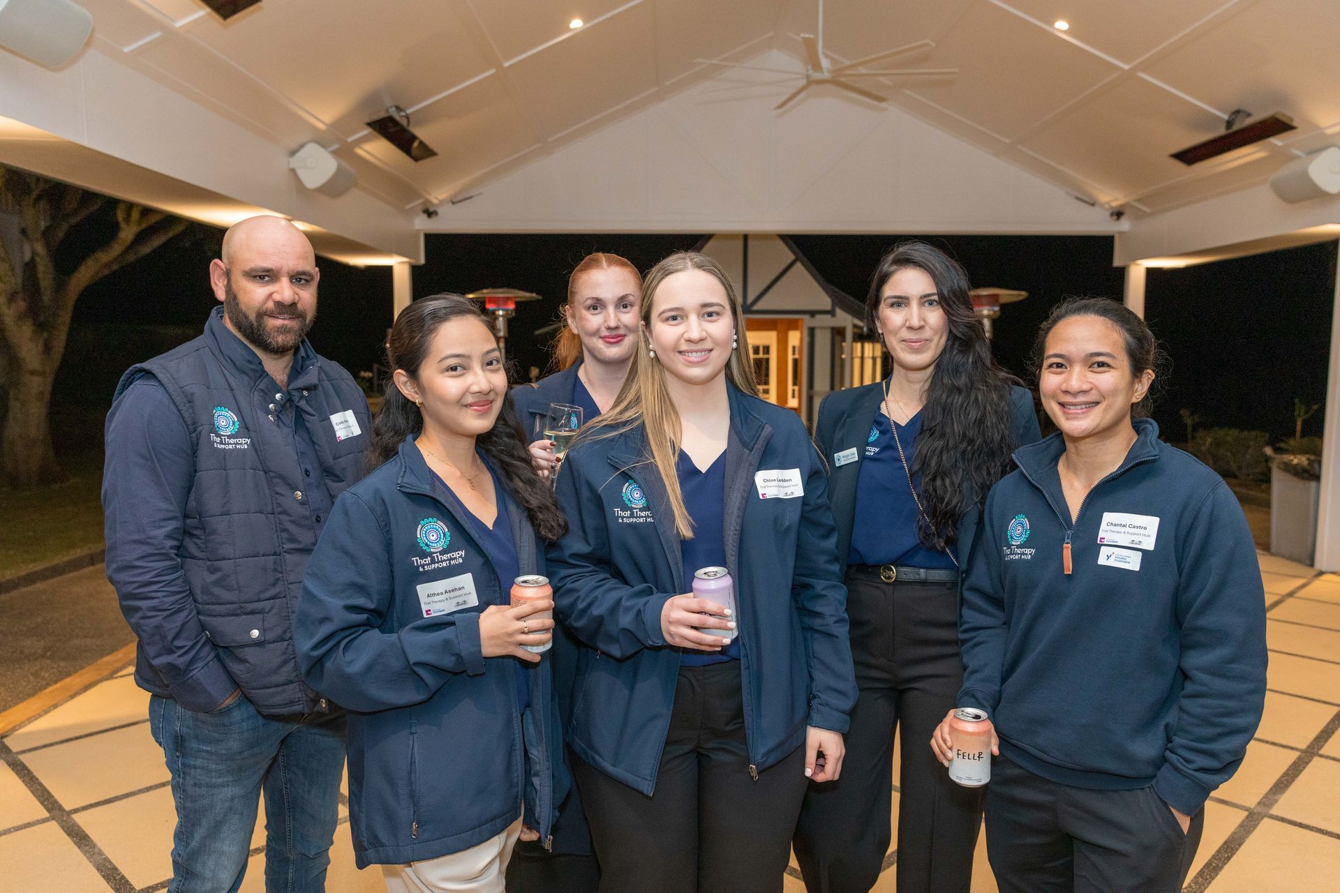 Group of six people, wearing matching navy jackets and name tags, smiling at an outdoor event under a covered patio.