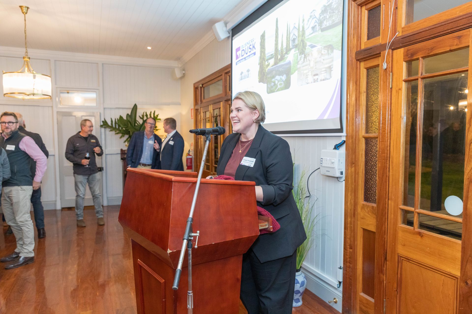 Woman speaking at a wooden podium in a room. A projector screen displays a presentation. People stand in the background.