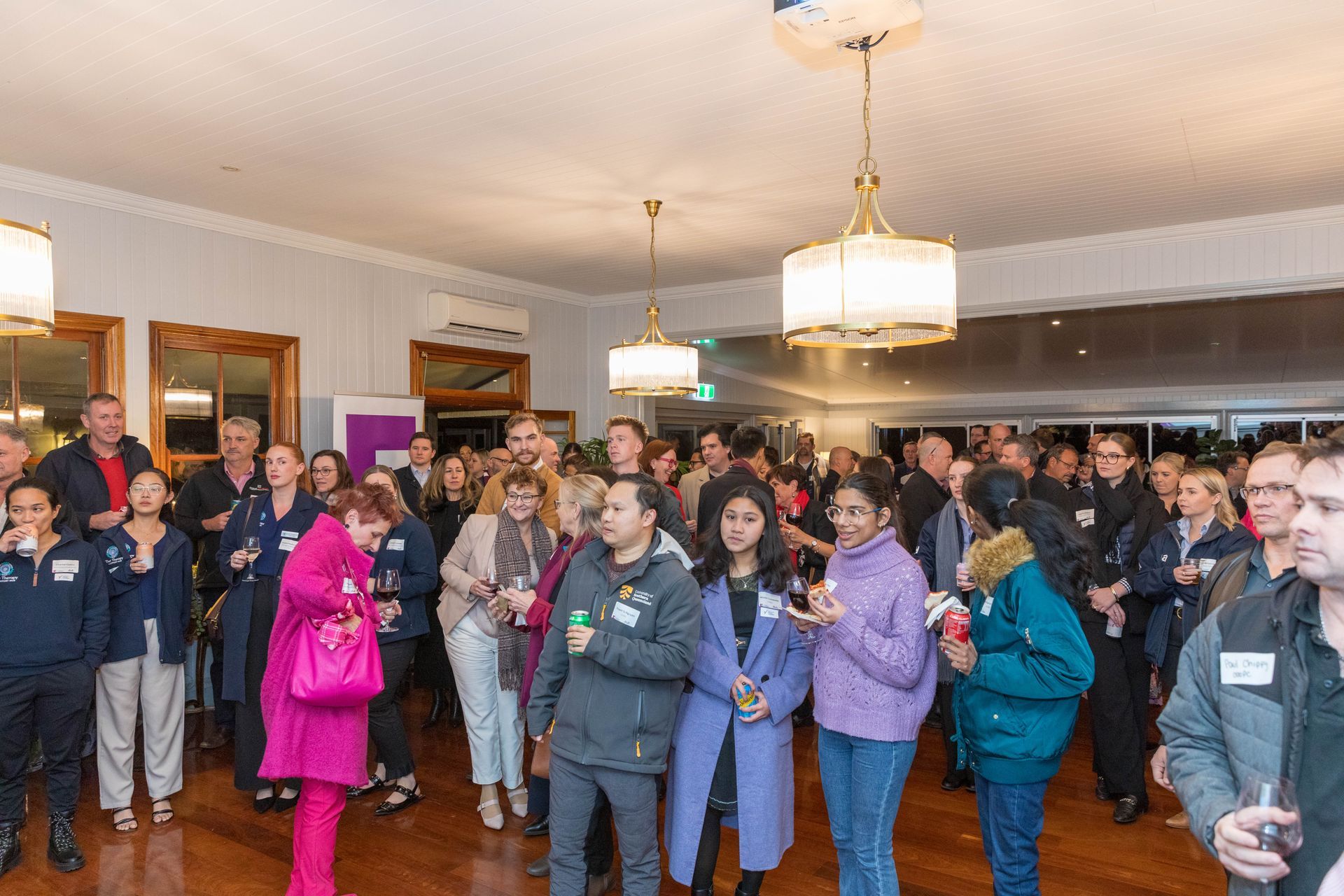 Crowd of diverse people in a well-lit room, possibly at an event. Some are talking and holding drinks.