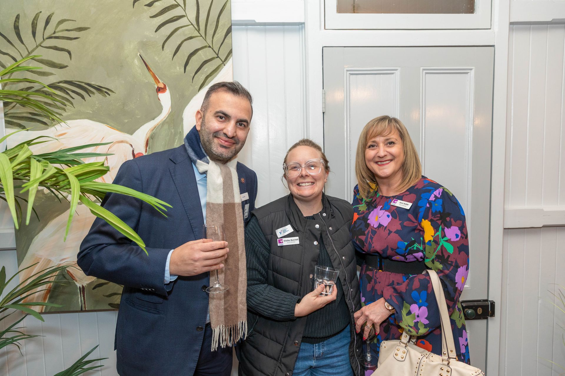 Three people smiling for a photo: a man in a blue suit, a woman in a black vest holding a glass, and a woman in a floral dress.