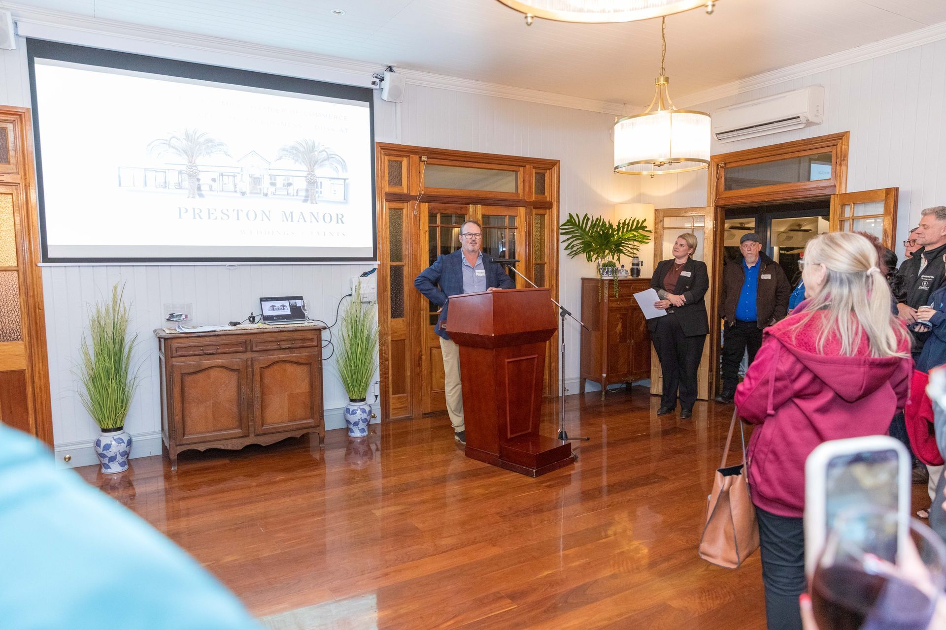 A man speaks at a podium in a wood-floored room. A projection screen displays a logo. People watch, some holding phones.