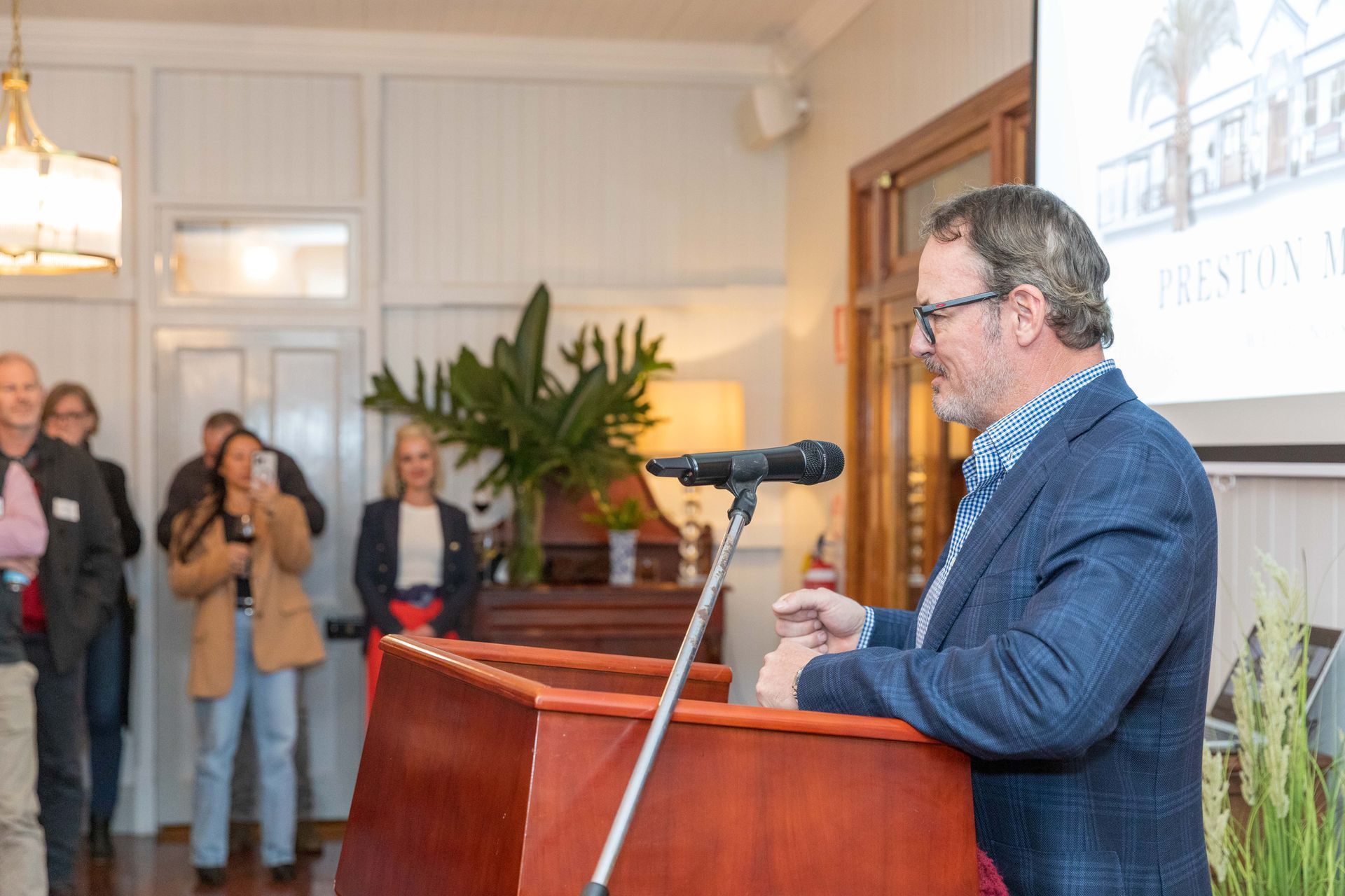 A man speaks from a podium at an event, with a crowd watching in a light-filled room.