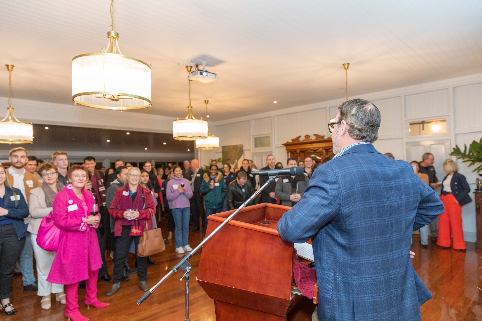 A man in a blue blazer speaks at a podium to a large audience in a well-lit room. People of various ages stand and listen.
