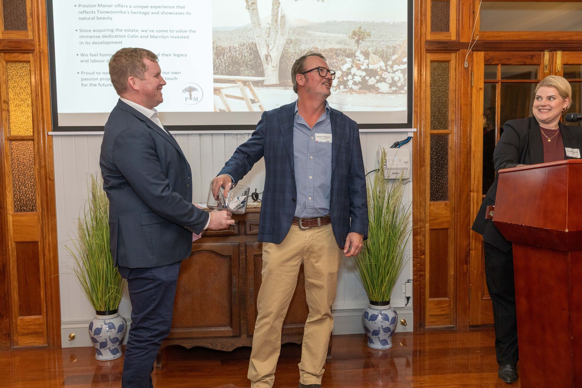 Two men shaking hands at an event; a woman stands at a podium on the right. The setting is a wood-paneled room.