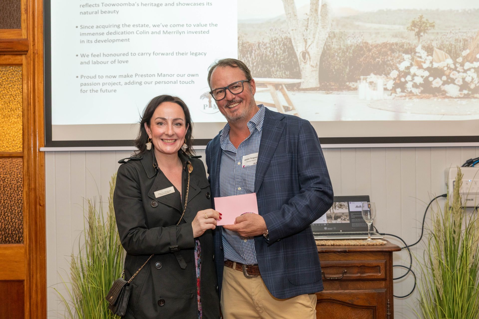 A woman in a black jacket presents a pink card to a smiling man in a blue blazer; they're in front of a slideshow screen and a table.