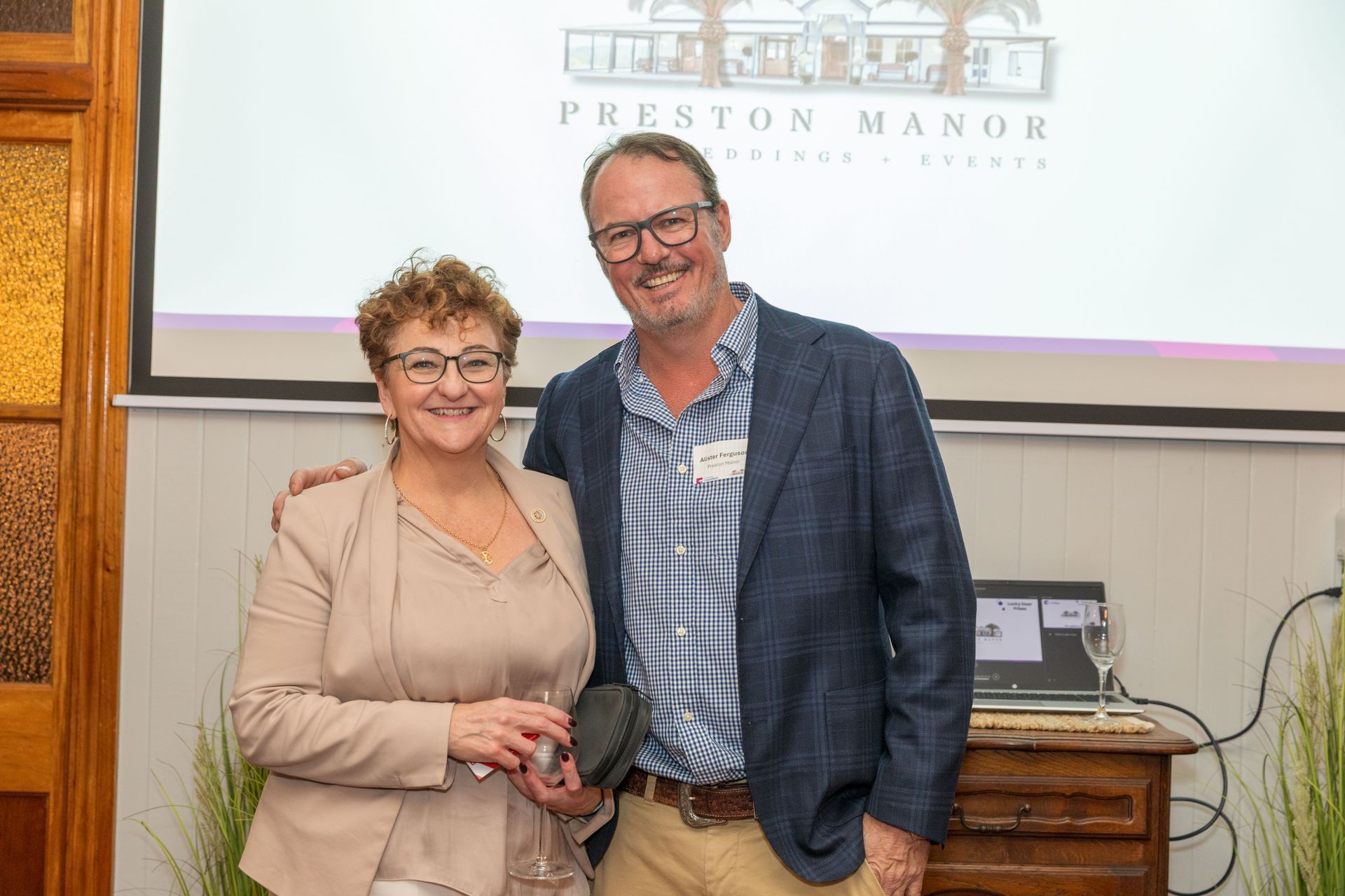 A woman in a beige suit and a man in a blue blazer pose together, smiling, in front of a Preston Manor sign.