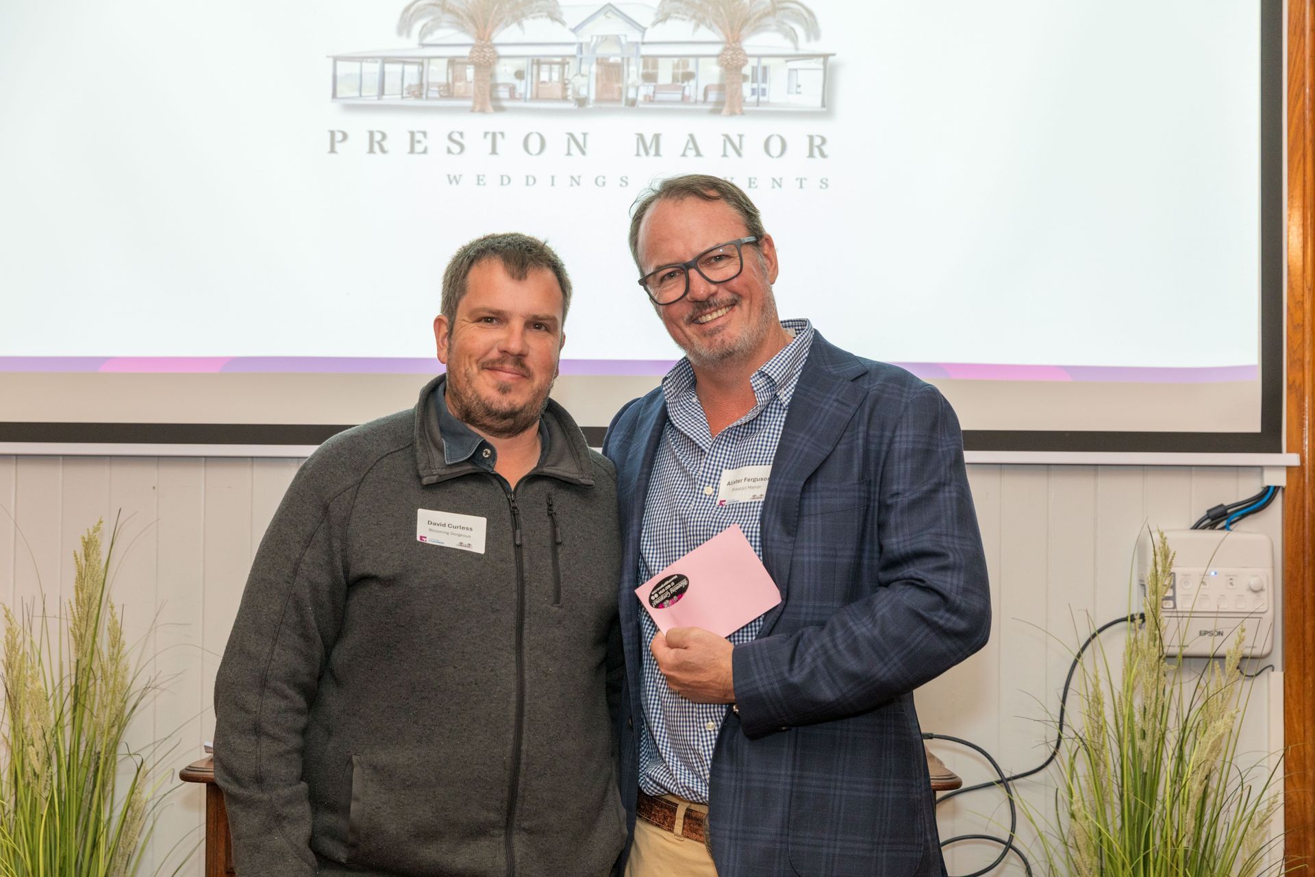 Two men smiling, posing together in front of a projector screen with a 