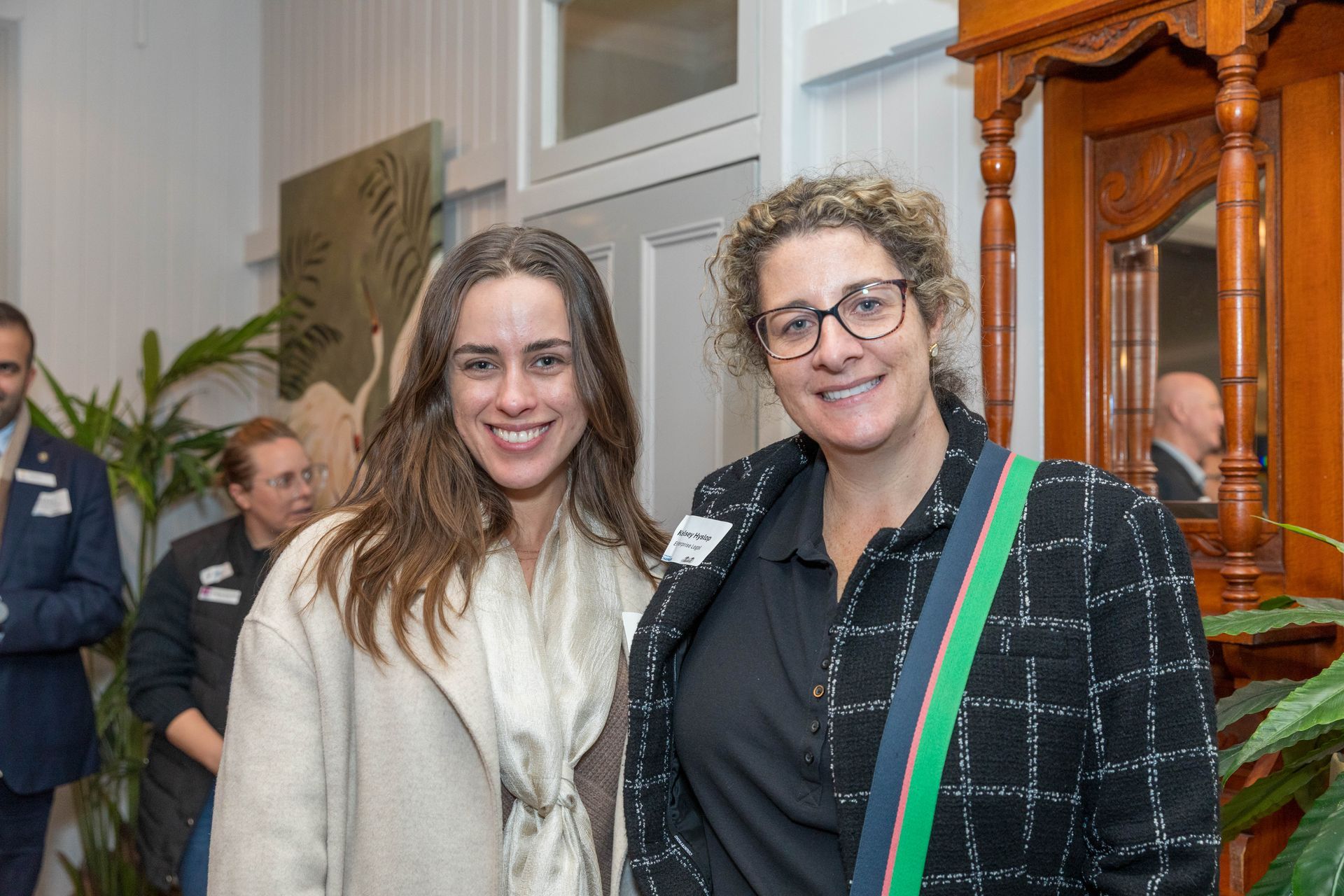 Two women smiling, standing inside a room. One wears a cream coat, the other a black and white checkered jacket and glasses.
