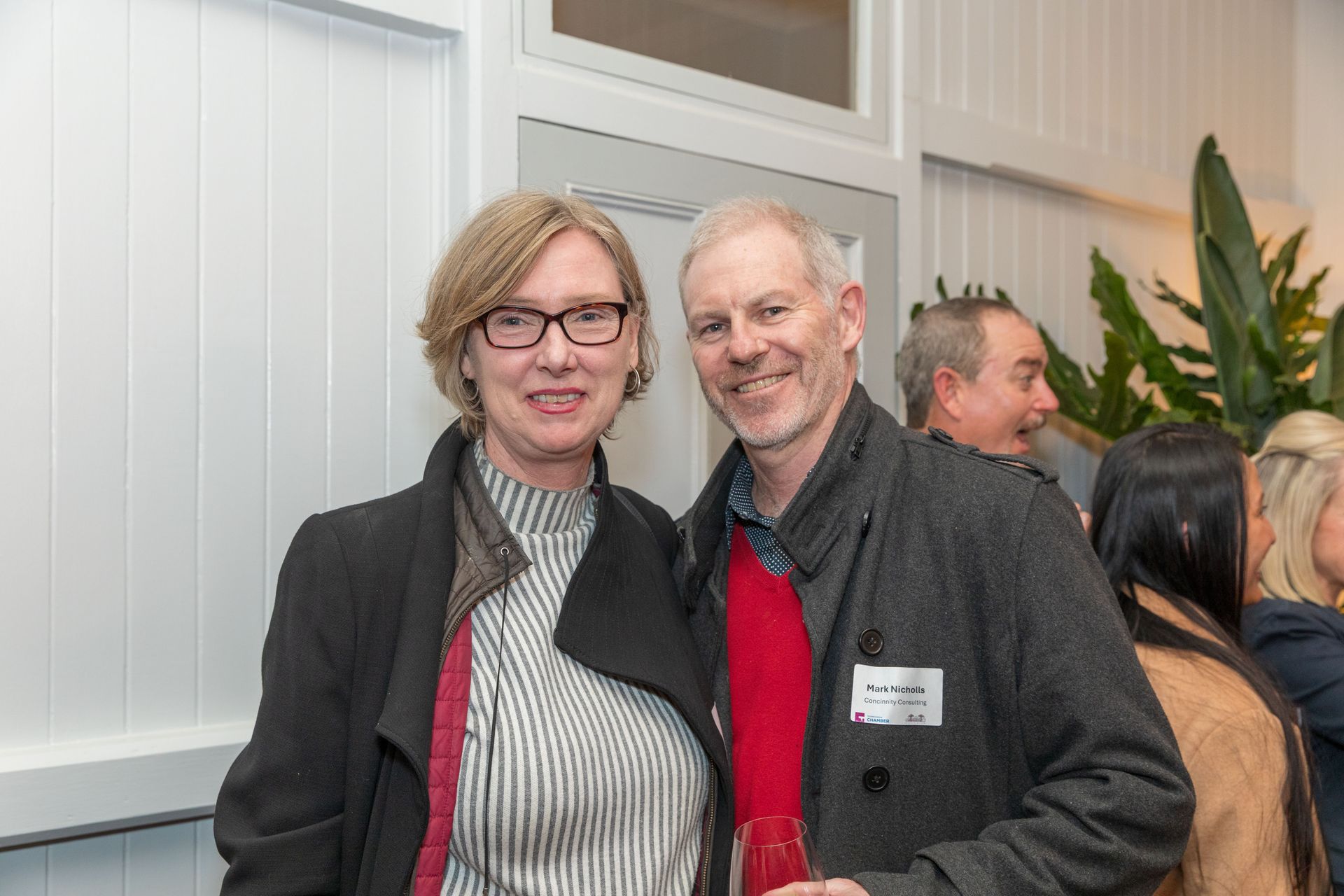 Two people smiling at a gathering. Woman with glasses and a coat, man with a red sweater. Other people in the background.
