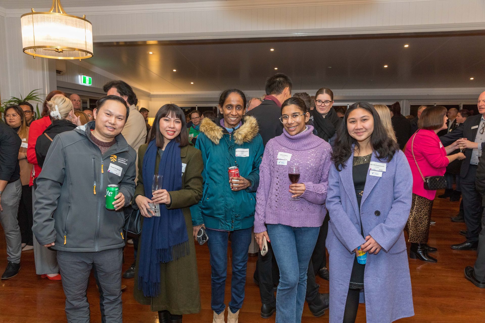Five people smiling at an event.  Four women and one man, all wearing coats, holding drinks, indoors with others in the background.