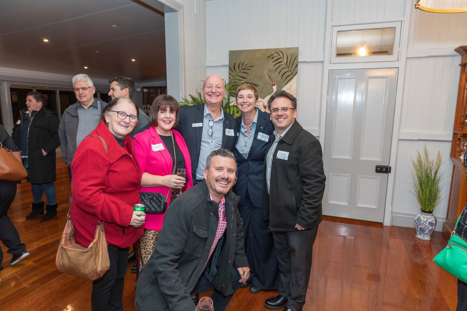 Group of people smiling for a photo at an event in a wood-floored room with white walls. Some are wearing name tags.