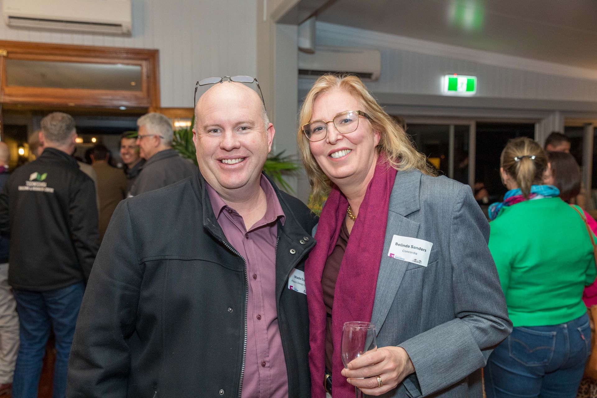 Two people smiling at a social gathering, man in jacket and burgundy shirt, woman in blazer and scarf, holding a glass.