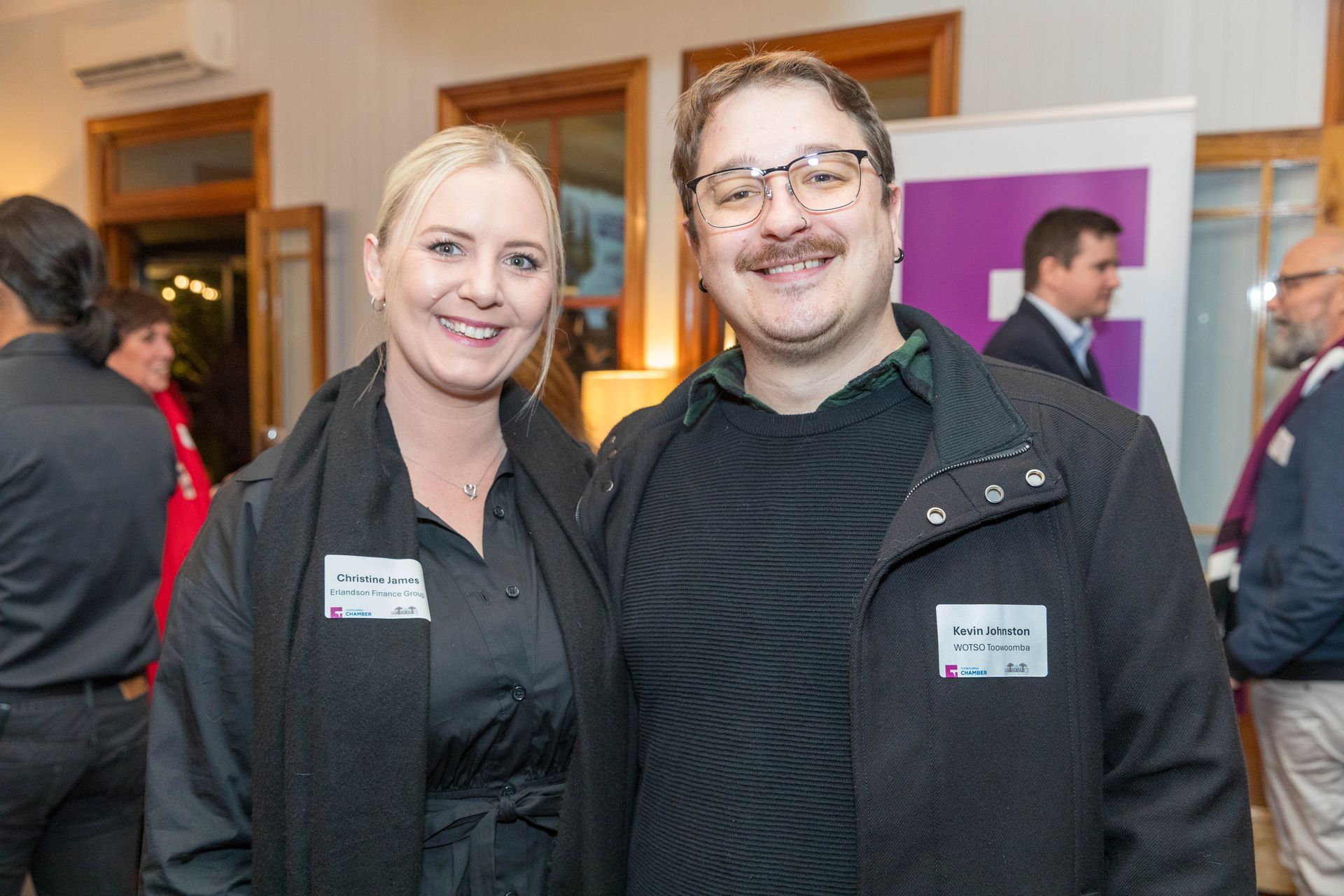 Two people smile at an event. A blonde woman wears black, and a man with glasses has a dark jacket. They are indoors.