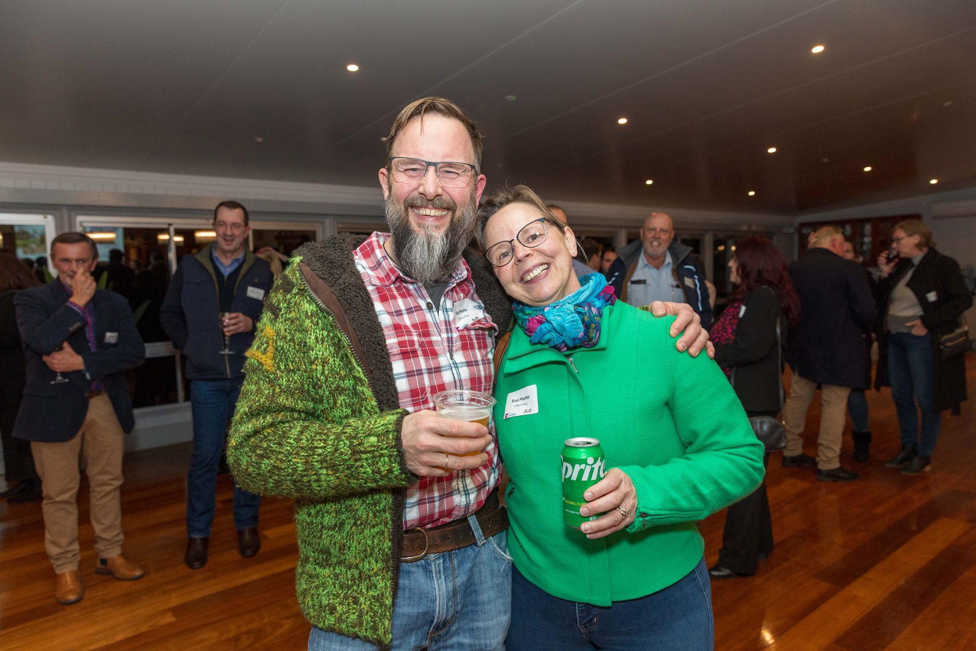 A smiling couple poses at an event. The man wears a red plaid shirt and green jacket, the woman a green jacket, both holding drinks.