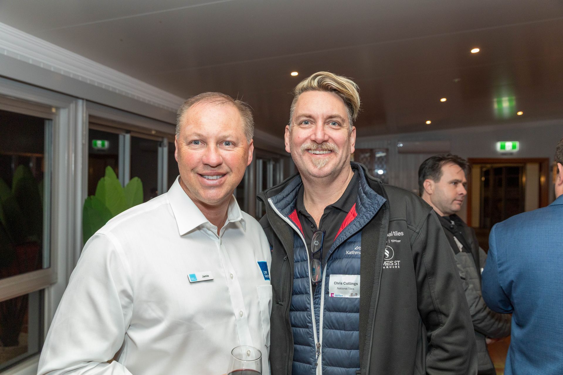 Two men smiling at an event. One in a white shirt, the other in a black jacket with blonde-tipped hair.