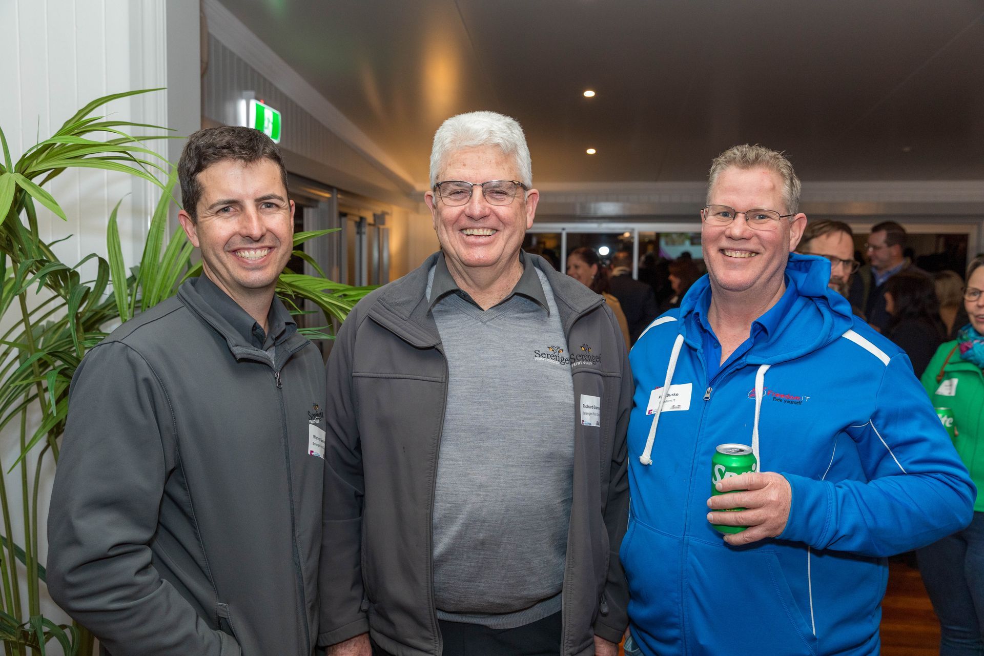 Three men smiling at an event; one on the right holds a soda can, others are wearing jackets.