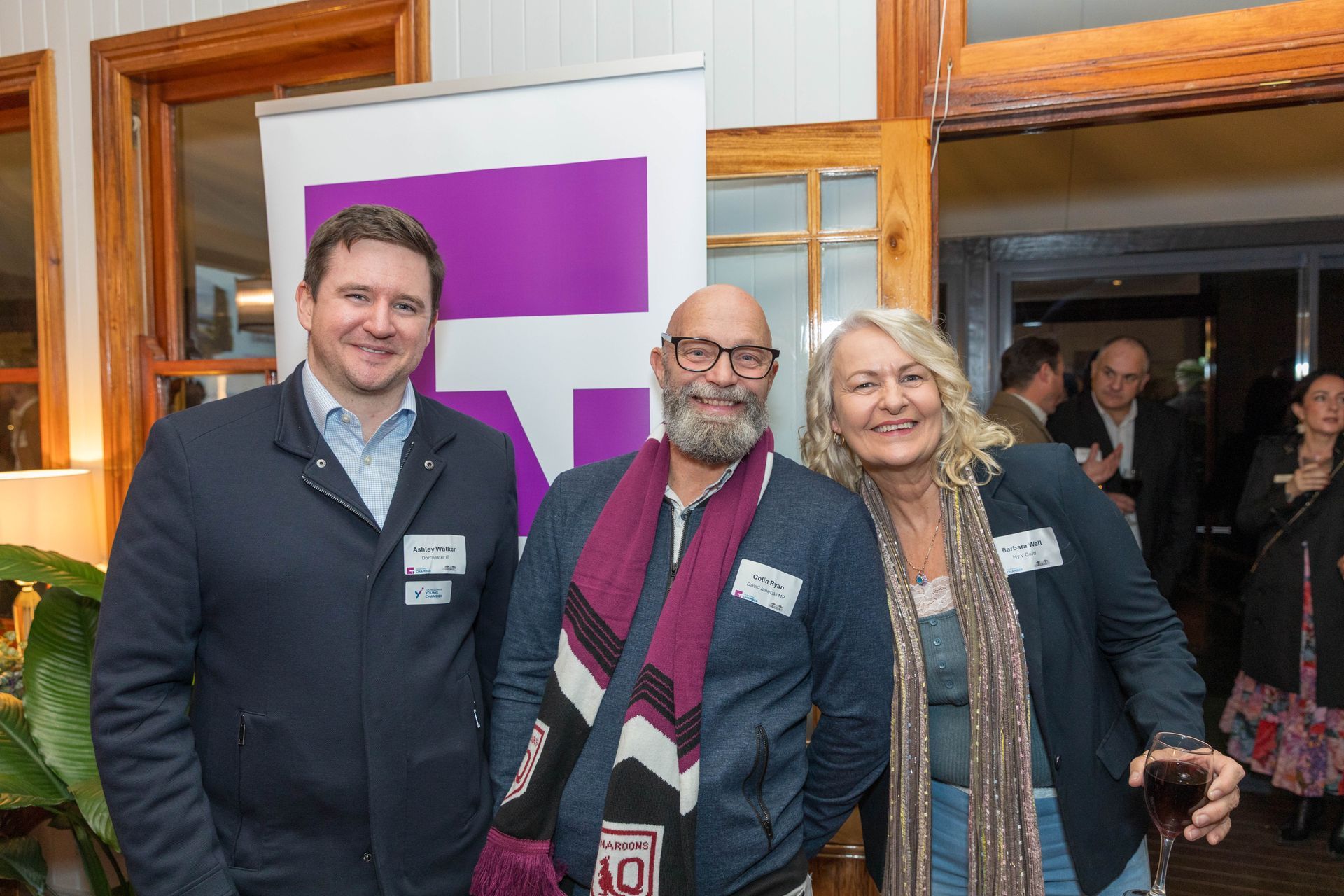 Three people smiling at an event, standing in front of a purple banner. Man on left wears a blazer; the middle man sports a scarf; the woman has blonde hair and a blazer, holding a wine glass.